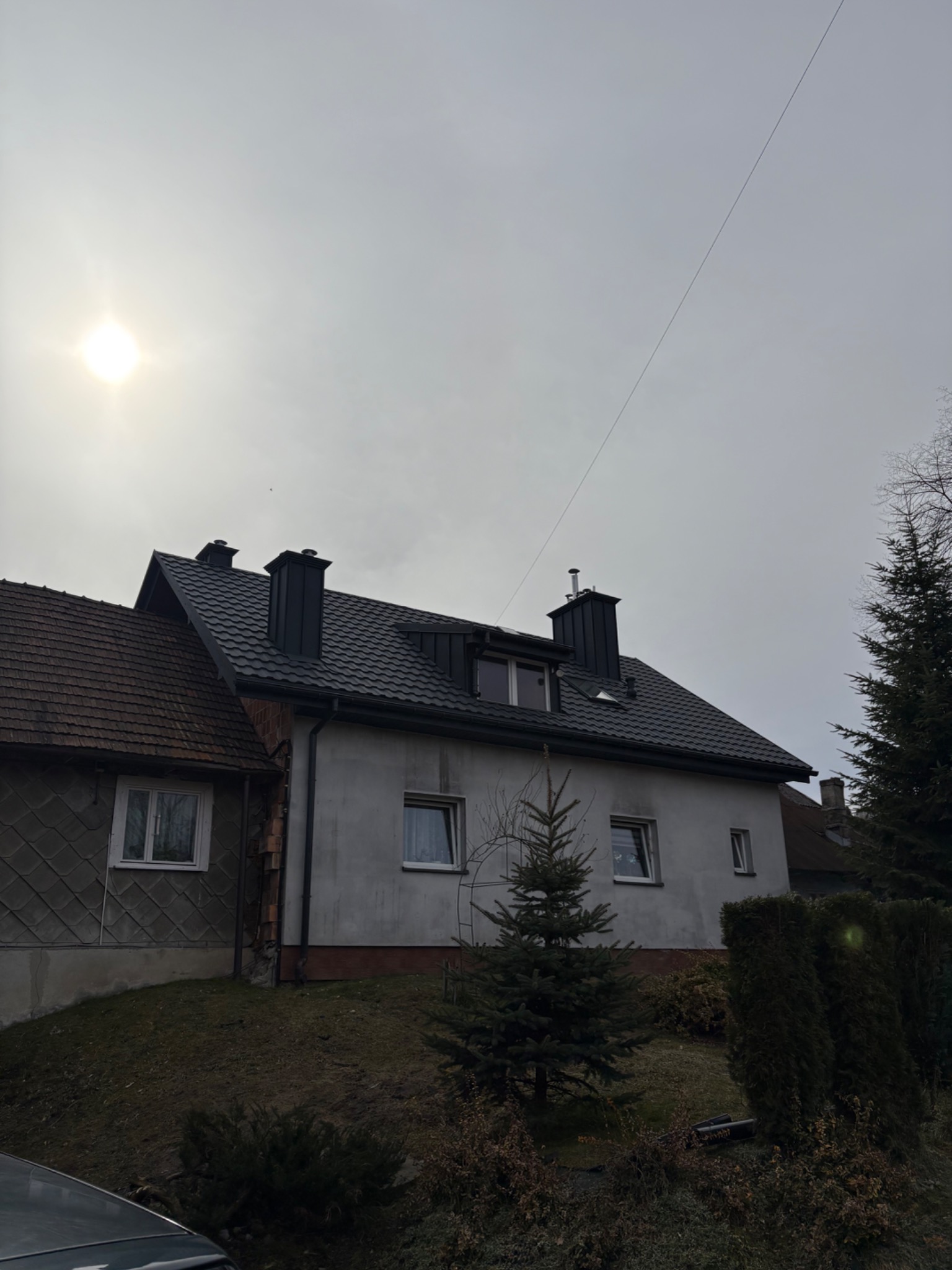 Dom z nowym, ciemnym dachem i kominami w pochmurny dzień. Widoczny fragment starego dachu sąsiedniego budynku. Na pierwszym planie iglaki i fragment samochodu.