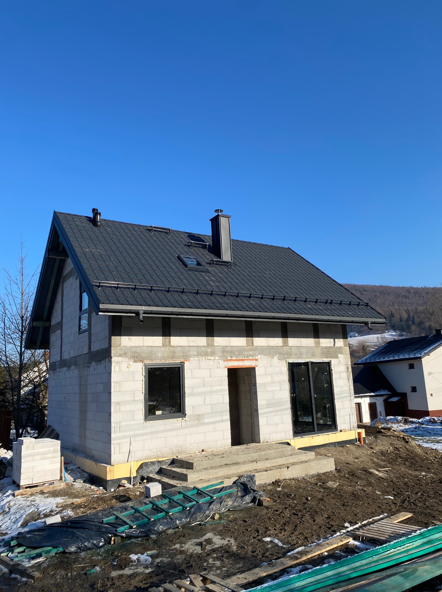Dom w stanie surowym zamkniętym z czarnym dachem na tle błękitnego nieba. Widoczne bloczki, okna, komin i częściowo ocieplenie. Wokół resztki śniegu i narzędzia.