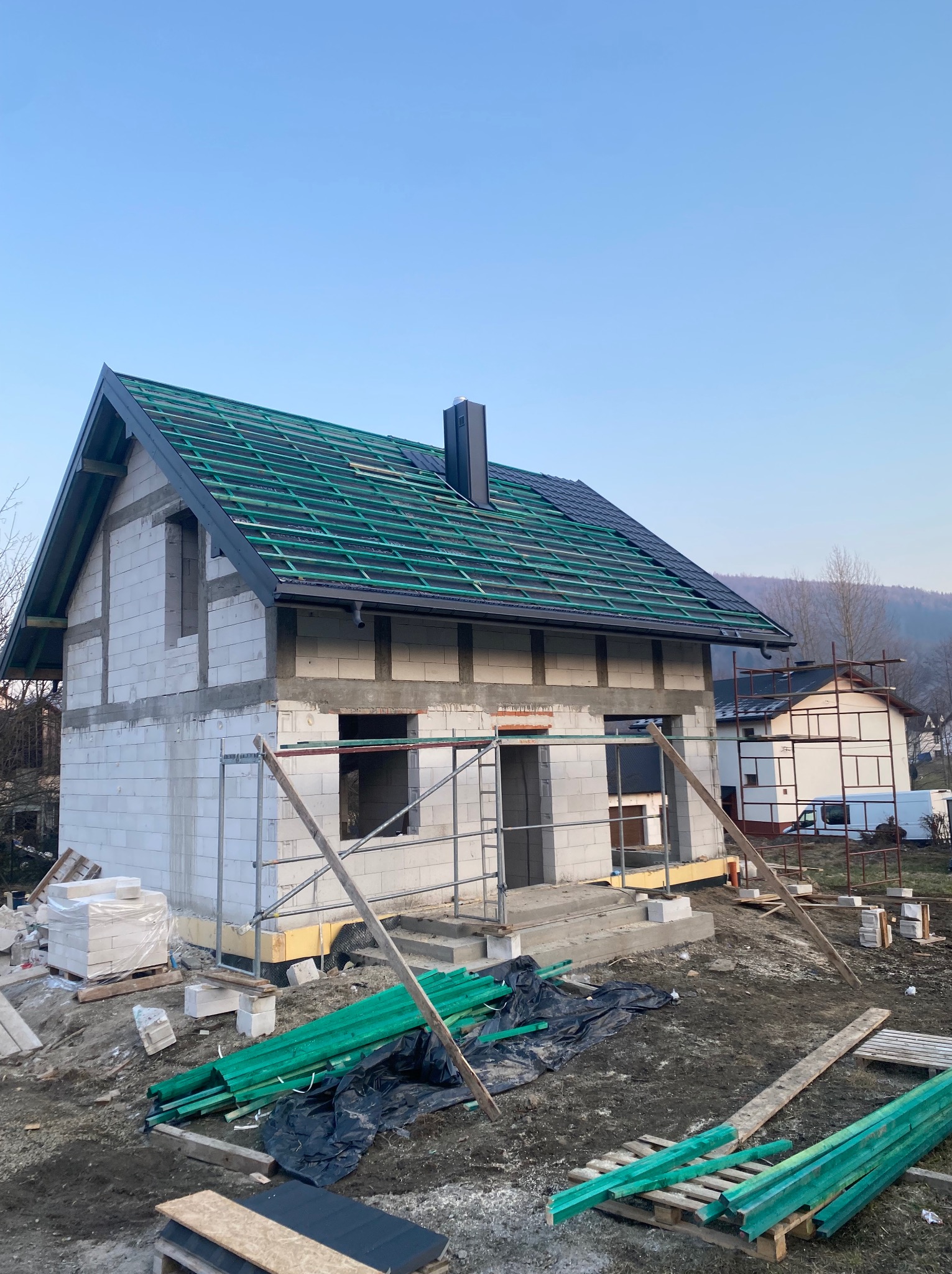 Budowa domu jednorodzinnego: surowy stan z widoczną konstrukcją dachu z łatami w kolorze zielonym i czarną blachą, na tle błękitnego nieba.