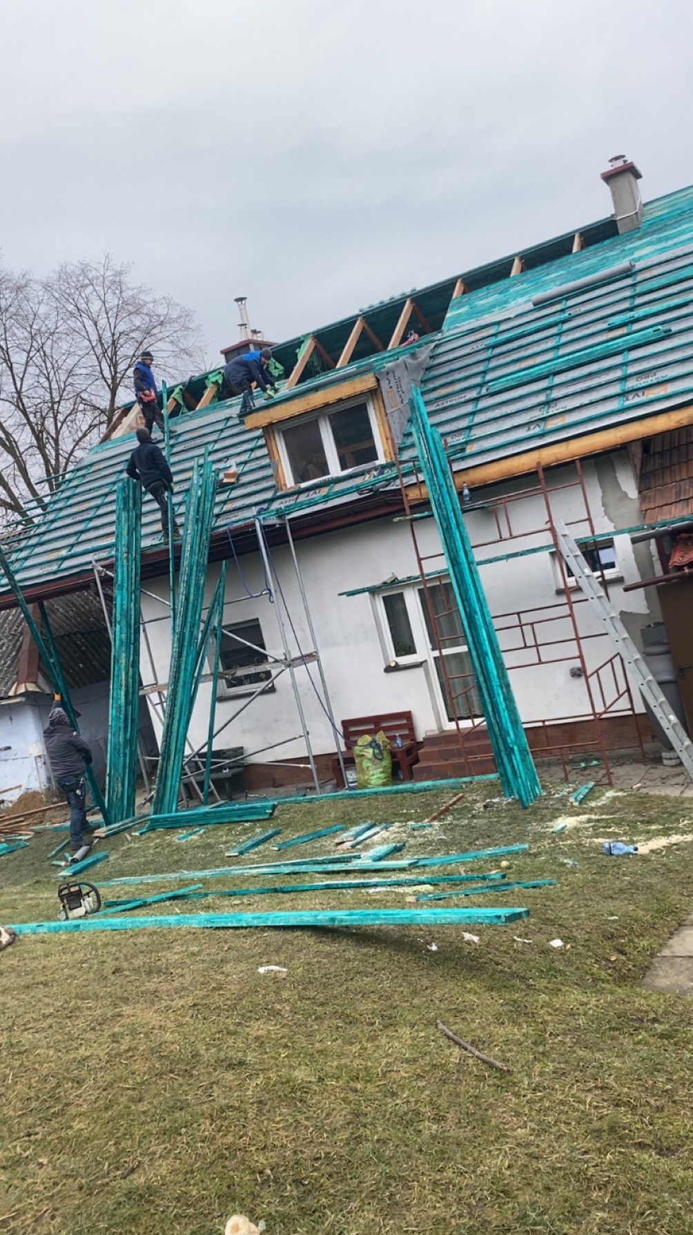 Prace dekarskie na dwuspadowym dachu domu jednorodzinnego. Widoczni dekarze na rusztowaniu, fragmenty desek i narzędzia na trawniku. Dach pokryty zieloną membraną.