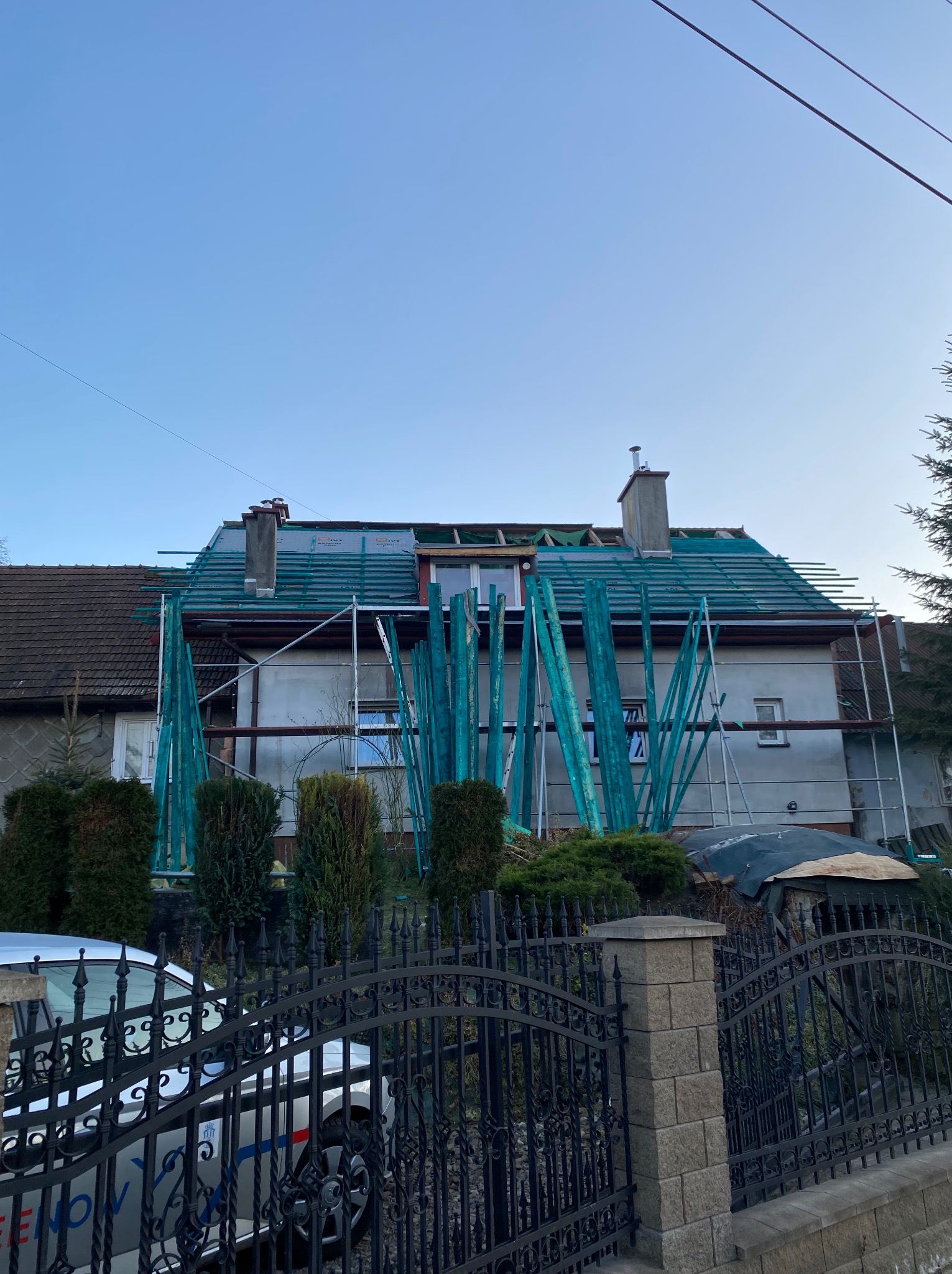 Dom jednorodzinny w trakcie prac dekarskich; widoczne rusztowania i częściowo położone zielone pokrycie dachu. Ujęcie zza ozdobnego, metalowego ogrodzenia.