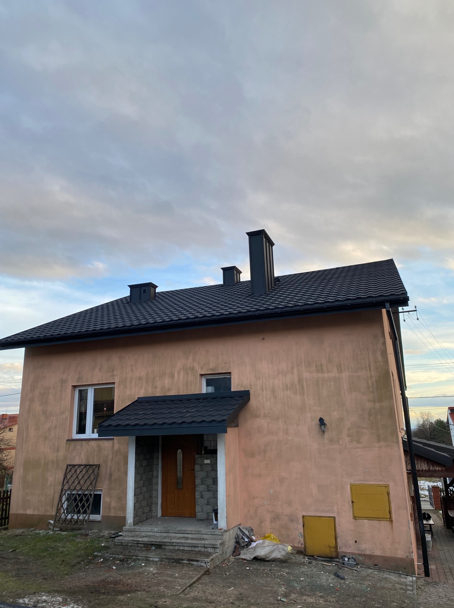 Dom z nowym, ciemnym dachem z dachówki, widocznymi kominami i fragmentem elewacji w odcieniach beżu. Ujęcie z dołu, pochmurne niebo w tle.