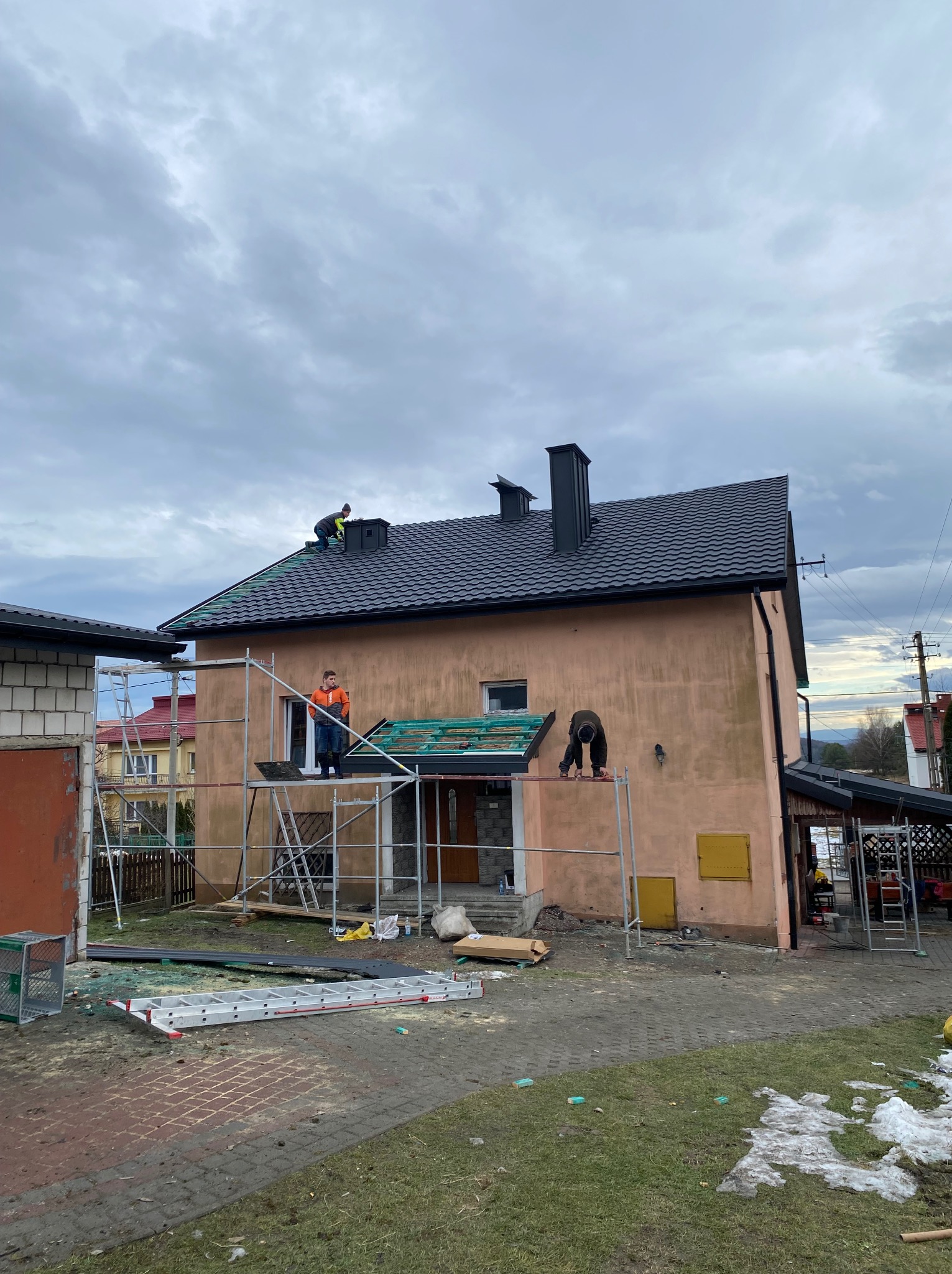 Montaż dachu na dwupiętrowym domu. Ekipa dekarzy na rusztowaniu układa czarne dachówki. Widoczne elementy konstrukcji dachu i narzędzia pracy. Pochmurne niebo w tle.