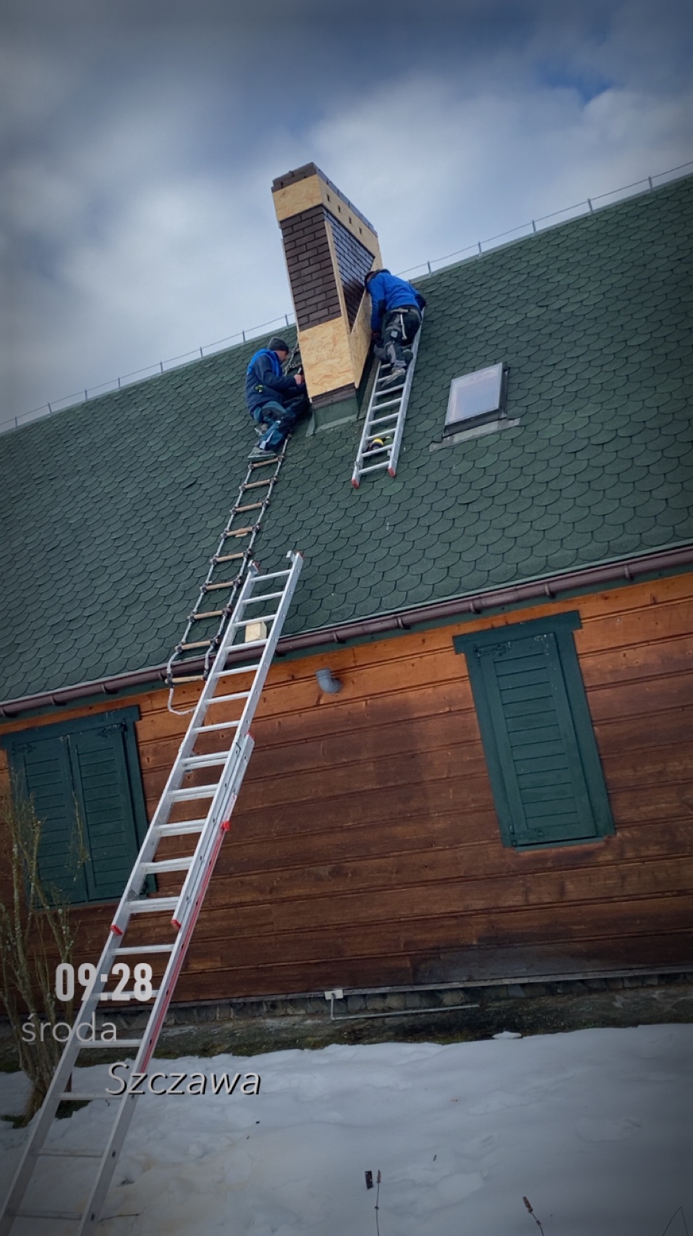 Dwóch dekarzy na drabinach montuje obudowę komina na dachu domu z zielonym gontem bitumicznym. Praca w toku, zimowa sceneria.