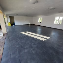 Grzegorz Siembida-Wykończenia wnętrz - Wnętrze przestronnego, jasnego pomieszczenia z nową, ciemną podłogą z płytek imitujących drewno, widoczne okna z białymi ramami, białe ściany i sufit z zamontowanymi lampami, na zewnątrz widoczny...