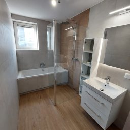 Grzegorz Siembida-Wykończenia wnętrz - Nowoczesna, jasna łazienka po remoncie w Ulanowie. Widoczna wanna z parawanem nawannowym, umywalka z szafką i lustrem z oświetleniem LED, drewnopodobna podłoga i częściowo wykończona drewnem ściana...