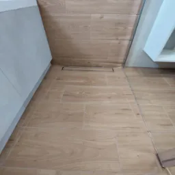 Ułożone płytki podłogowe i ścienne imitujące drewno, z widocznym odpływem liniowym prysznicowym i fragmentem kabiny prysznicowej oraz szafki pod umywalkę.