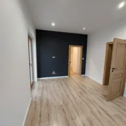 Wnętrze po remoncie: jasne panele podłogowe, białe ściany z elementami czarnej ściany, dwoje drewnianych drzwi z czarnymi klamkami, widoczne gniazdka elektryczne.