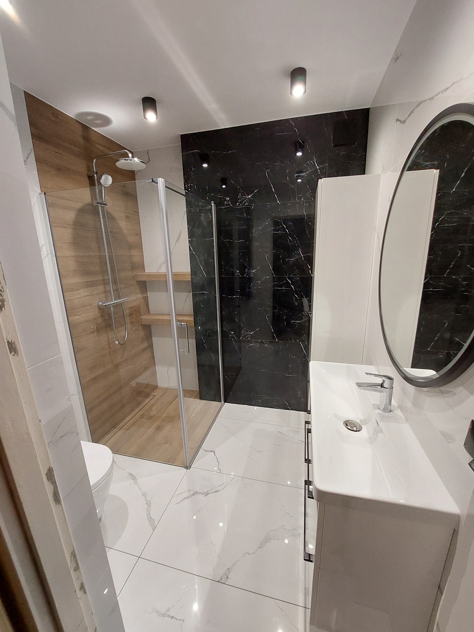 Nowoczesna łazienka z kabiną prysznicową typu walk-in z drewnopodobnymi płytkami, czarną ścianą z marmurowym wzorem, białą umywalką z szafką i owalnym lustrem w czarnej ramie.