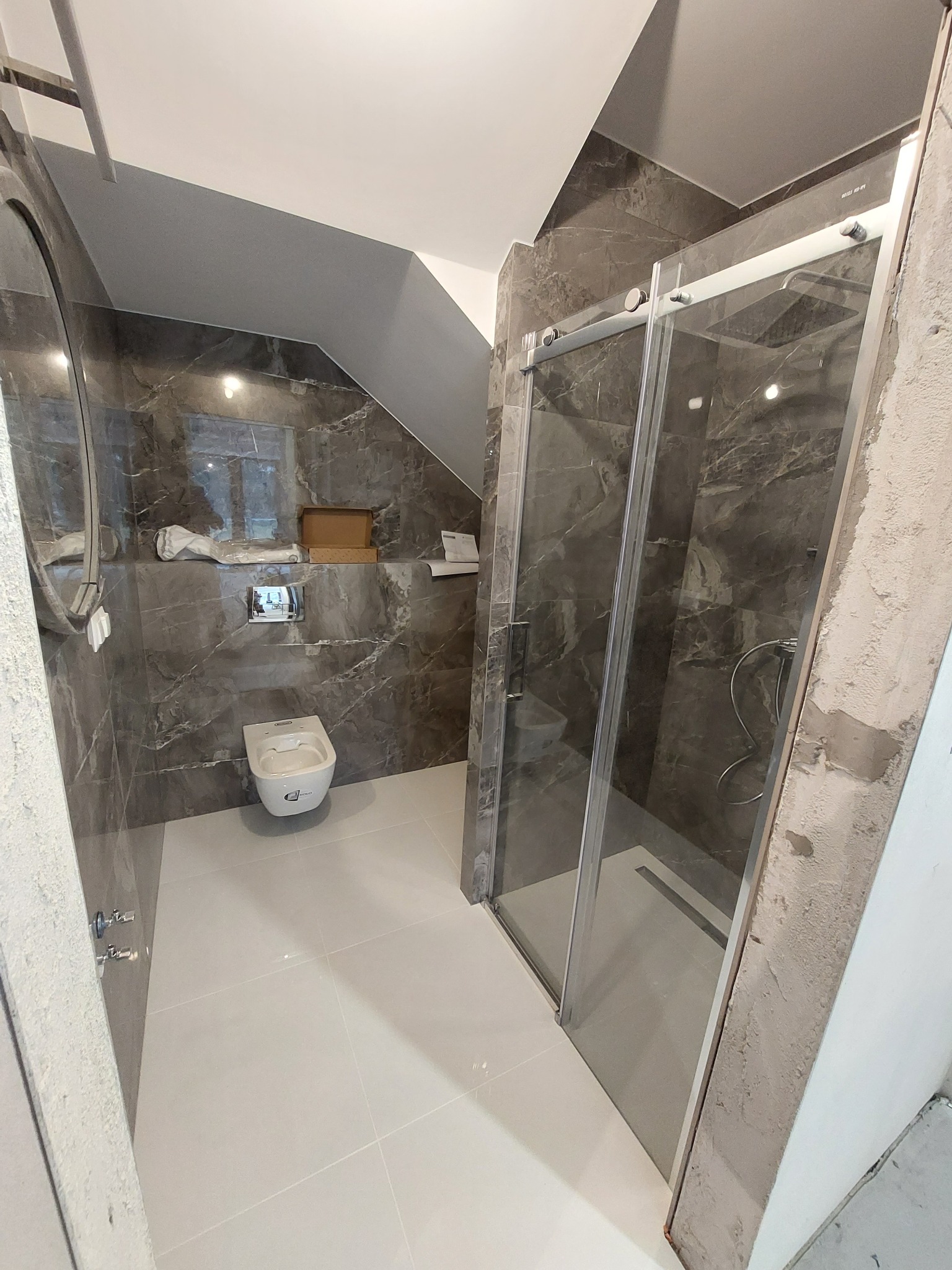 Ukośna łazienka z marmurowymi płytkami, białą ceramiką i kabiną prysznicową ze srebrnymi ramami, w trakcie montażu, widoczne niedokończone wykończenia ścian.