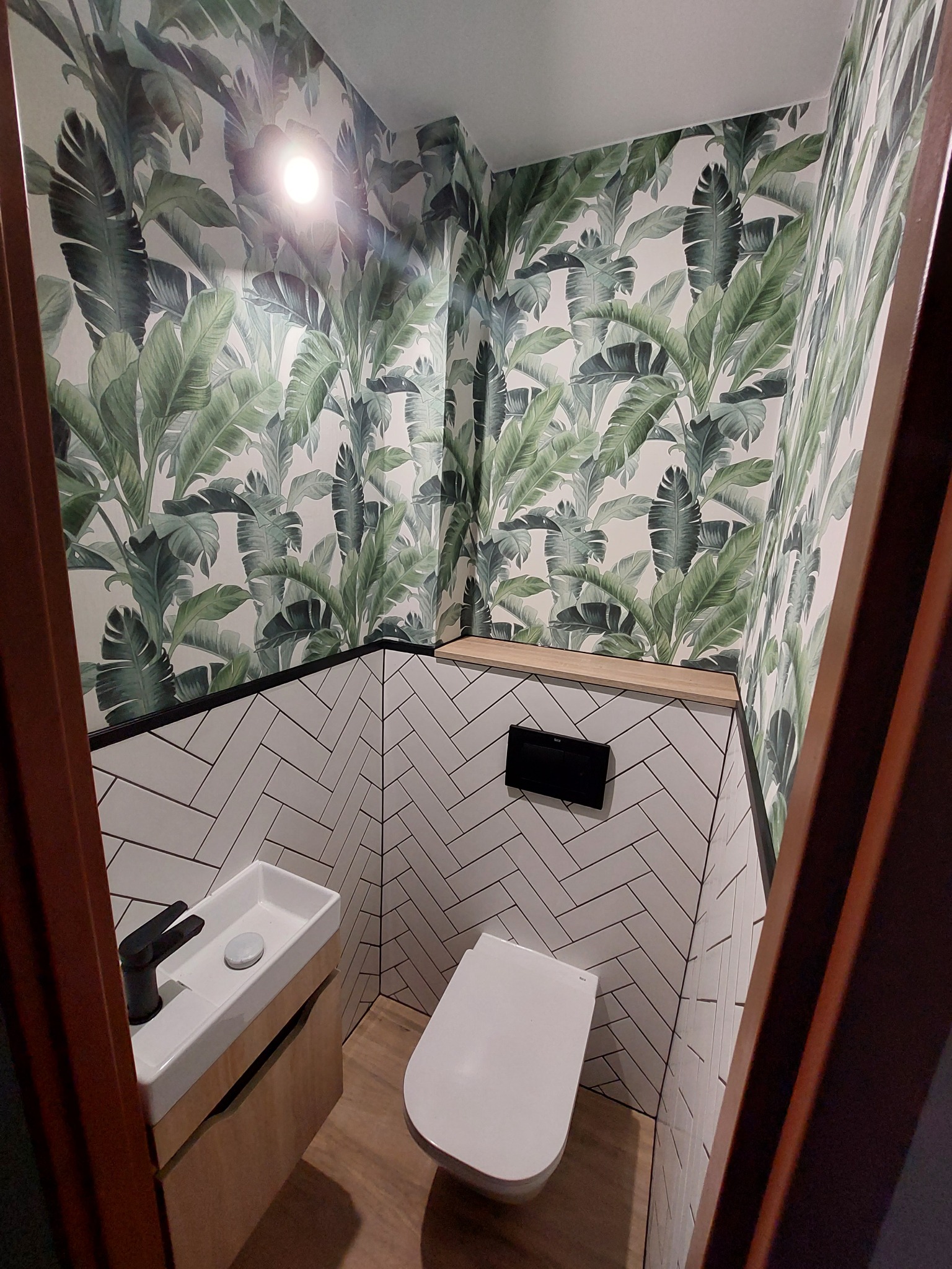 Nowoczesna, mała łazienka z toaletą podwieszaną, umywalką nablatową z czarną armaturą, białymi płytkami ułożonymi w jodełkę oraz tapetą w motywy tropikalnych liści.