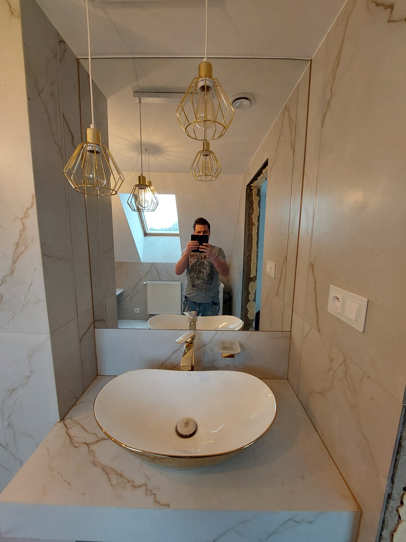 Nowoczesna łazienka z owalną umywalką nablatową z baterią w kolorze złota, marmurowe płytki na ścianach i suficie oraz geometryczne lampy wiszące z metalu w kolorze złota, widoczne odbicie osoby...
