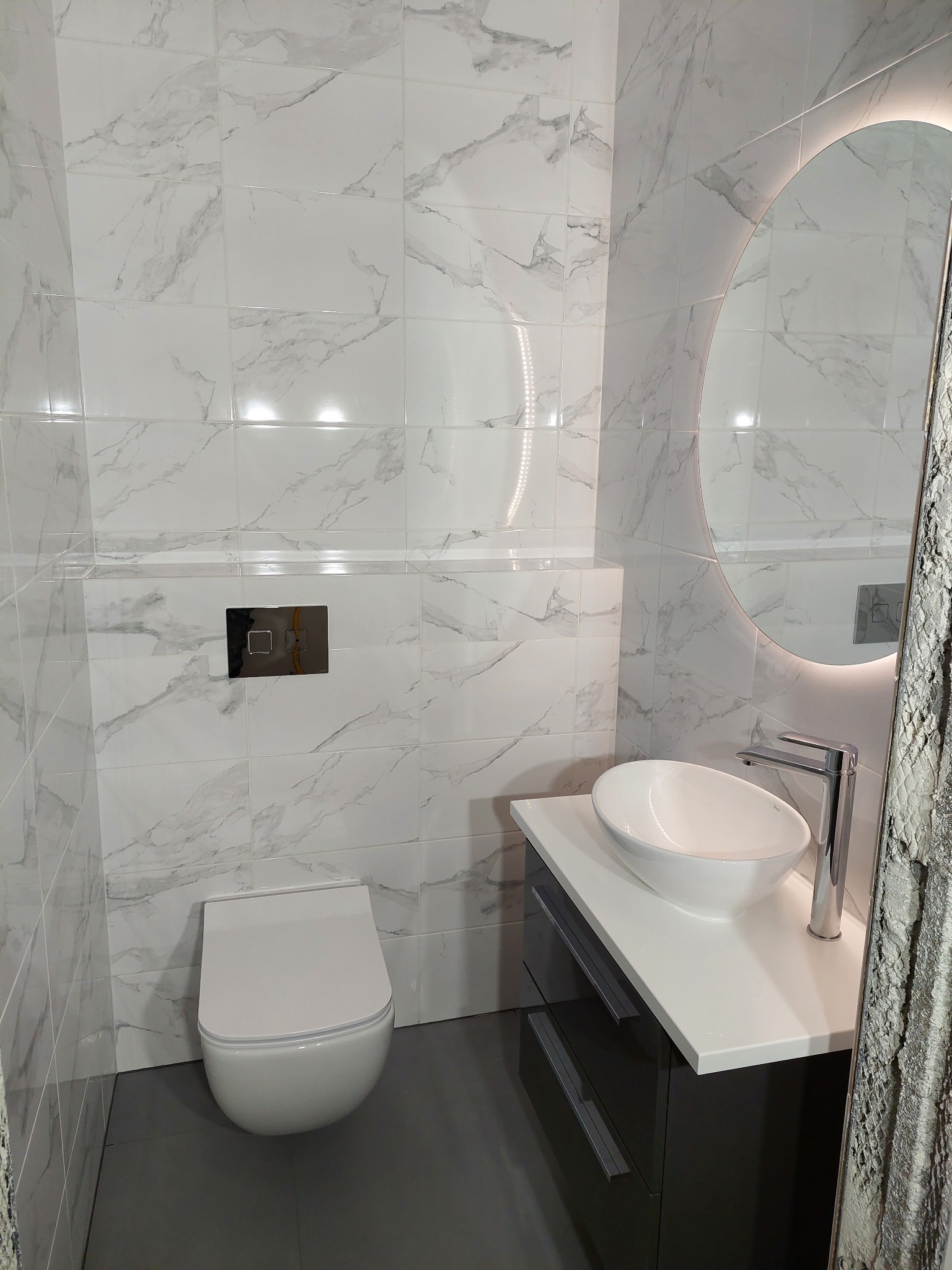 Nowoczesna łazienka z białymi marmurkowymi płytkami, owalnym lustrem z podświetleniem LED, wiszącą miską WC i umywalką nablatową na szafce z szufladami.