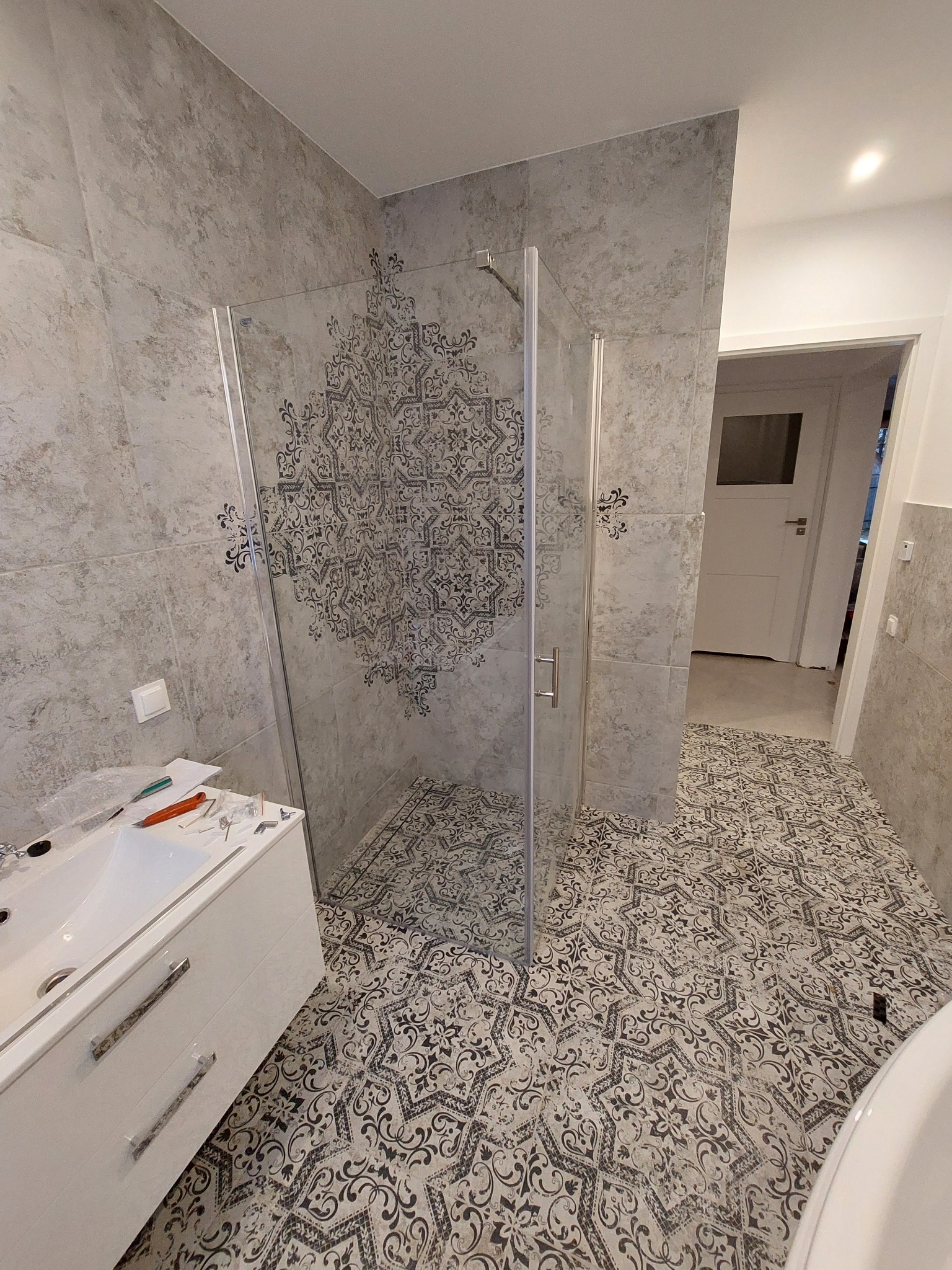 Wykończona łazienka z kabiną prysznicową ze szklanymi drzwiami, wzorzystymi płytkami podłogowymi i ściennymi, białą szafką pod umywalką oraz narzędziami na blacie.