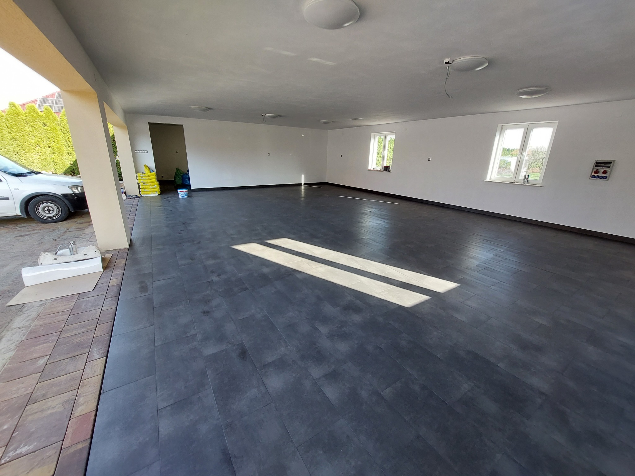 Wnętrze przestronnego, jasnego pomieszczenia z nową, ciemną podłogą z płytek imitujących drewno, widoczne okna z białymi ramami, białe ściany i sufit z zamontowanymi lampami, na zewnątrz widoczny...