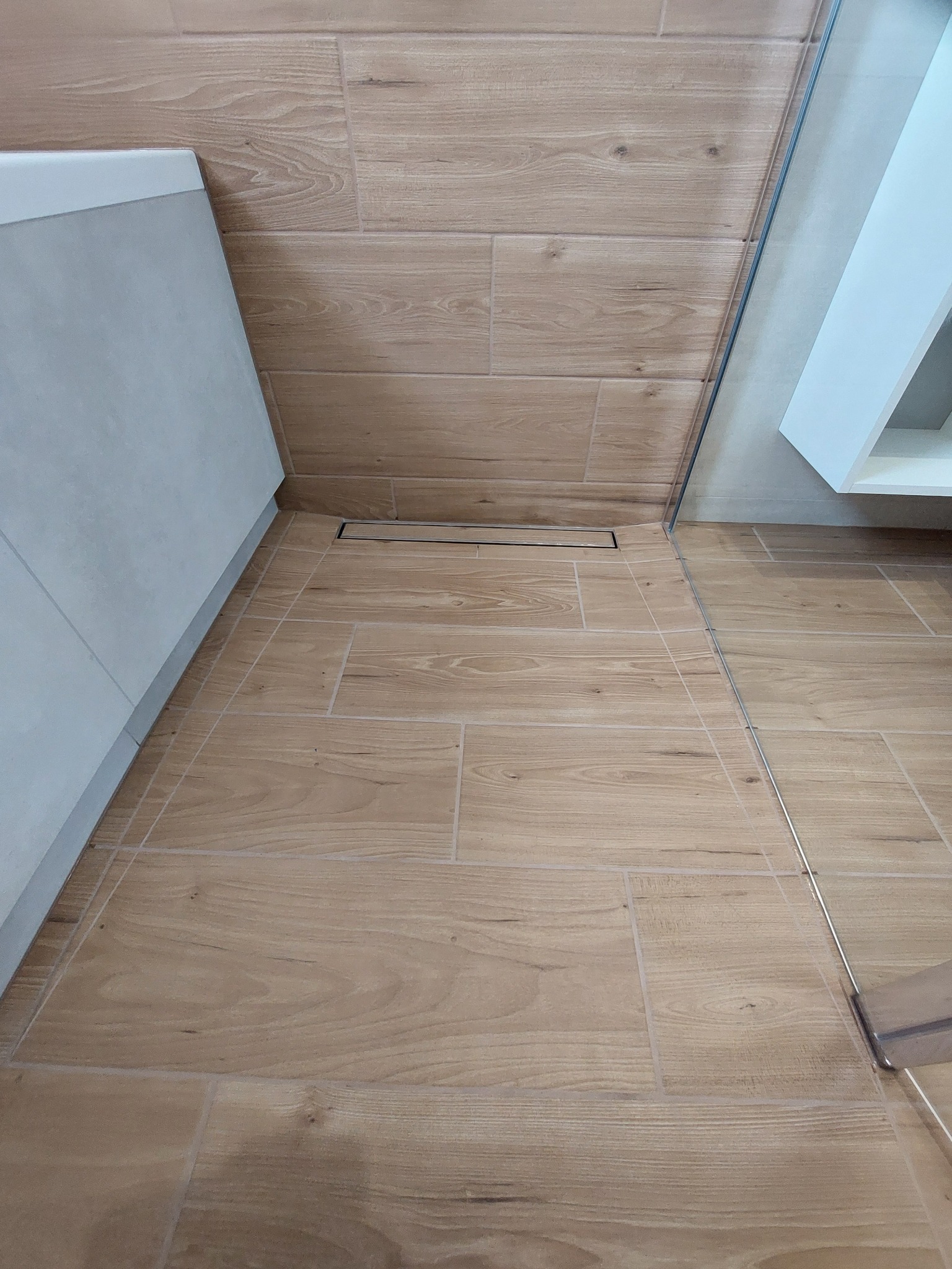 Ułożone płytki podłogowe i ścienne imitujące drewno, z widocznym odpływem liniowym prysznicowym i fragmentem kabiny prysznicowej oraz szafki pod umywalkę.