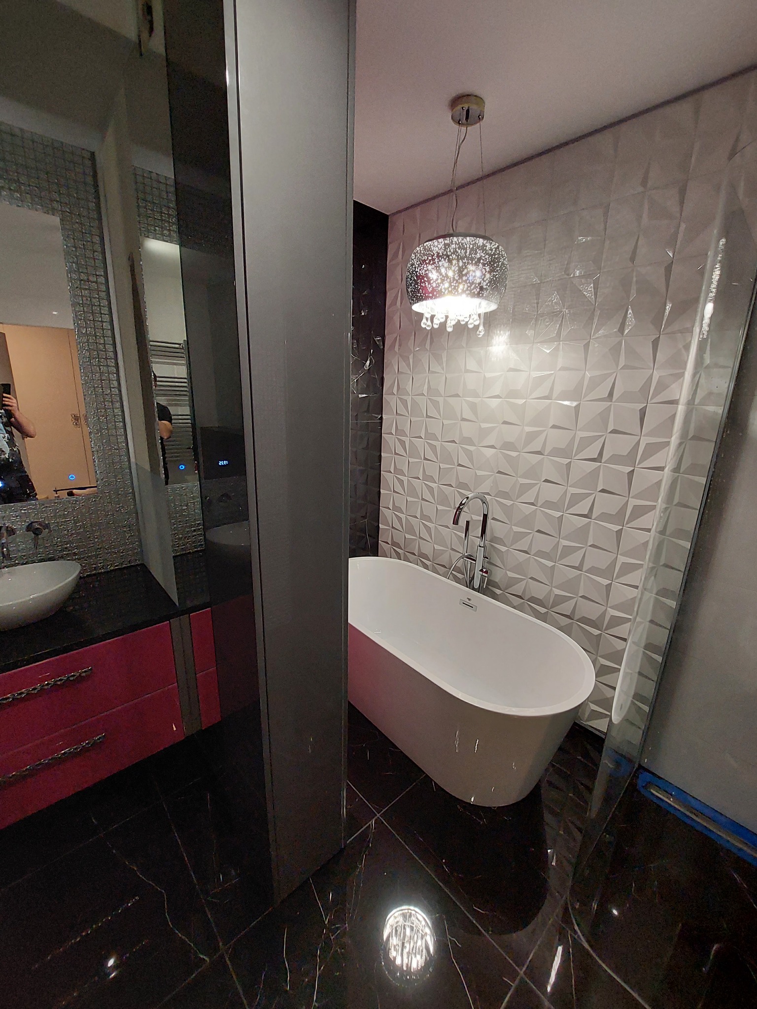 Nowoczesna łazienka z wolnostojącą wanną, trójwymiarową białą glazurą na ścianie, czarną marmurową podłogą i błyszczącym żyrandolem.