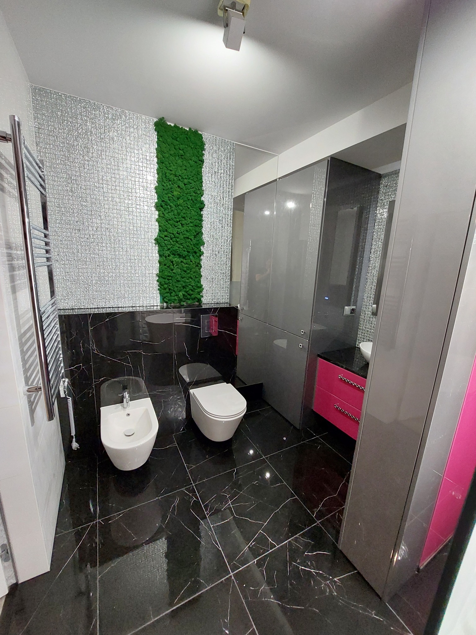 Nowoczesna łazienka z czarną marmurową podłogą i ścianą z mozaiki, pionowy panel z mchu, biała ceramika sanitarna i szafki w odcieniach szarości z różowymi akcentami.