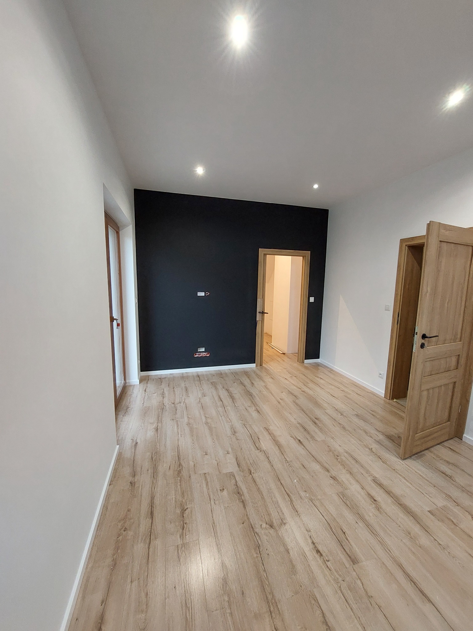 Wnętrze po remoncie: jasne panele podłogowe, białe ściany z elementami czarnej ściany, dwoje drewnianych drzwi z czarnymi klamkami, widoczne gniazdka elektryczne.