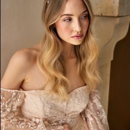 Katarzyna Tarnowiecka makeup artist & hair stylist - Portret kobiety z blond włosami w sukni z koronkowymi rękawami i ozdobną opaską, pozującej przy ścianie.