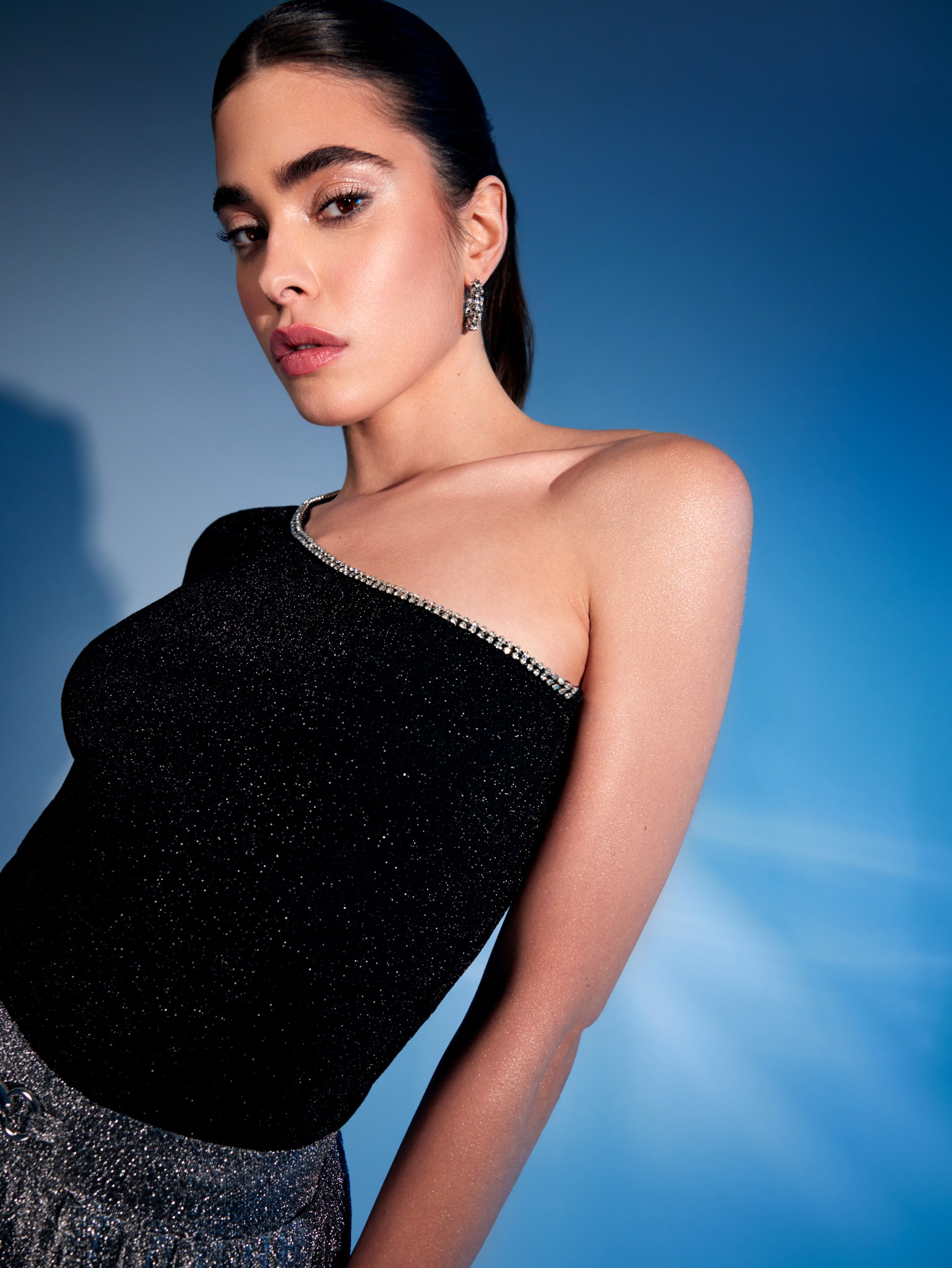 Portret kobiety z błyszczącym makijażem oczu i ust, w czarnej, cekinowej bluzce na tle gradientowego niebieskiego tła, z widocznymi kolczykami.