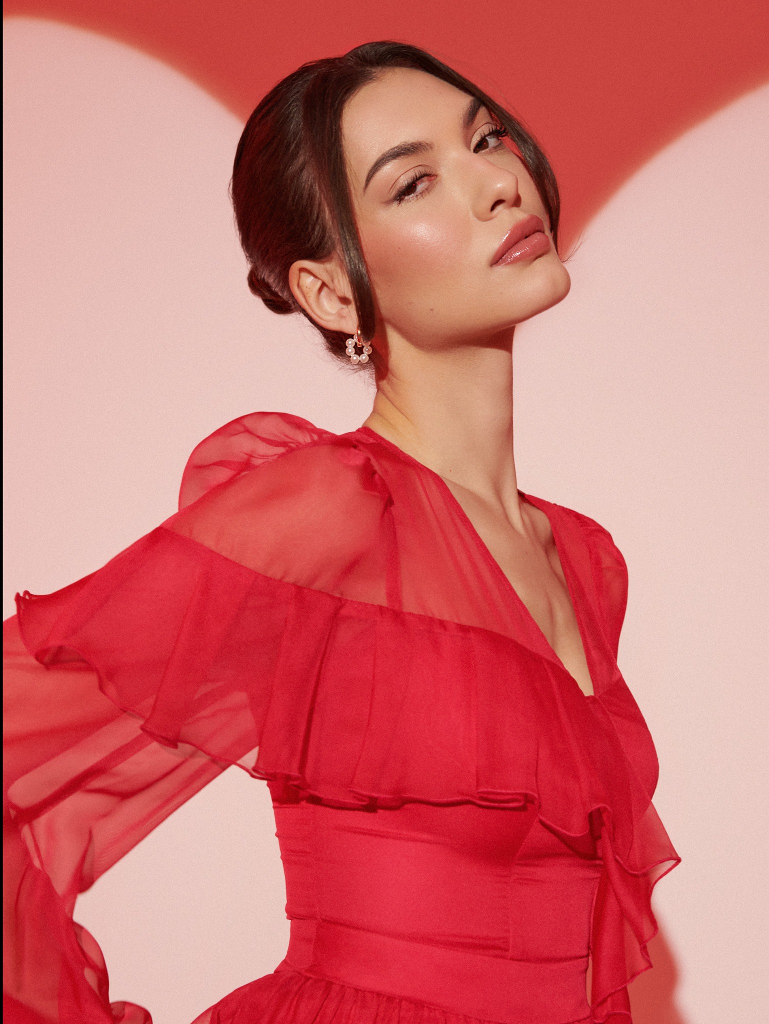 Portret kobiety z upiętymi włosami i delikatnym, błyszczącym makijażem, w czerwonej, zwiewnej sukience na tle różowego studia z efektem świetlnym.