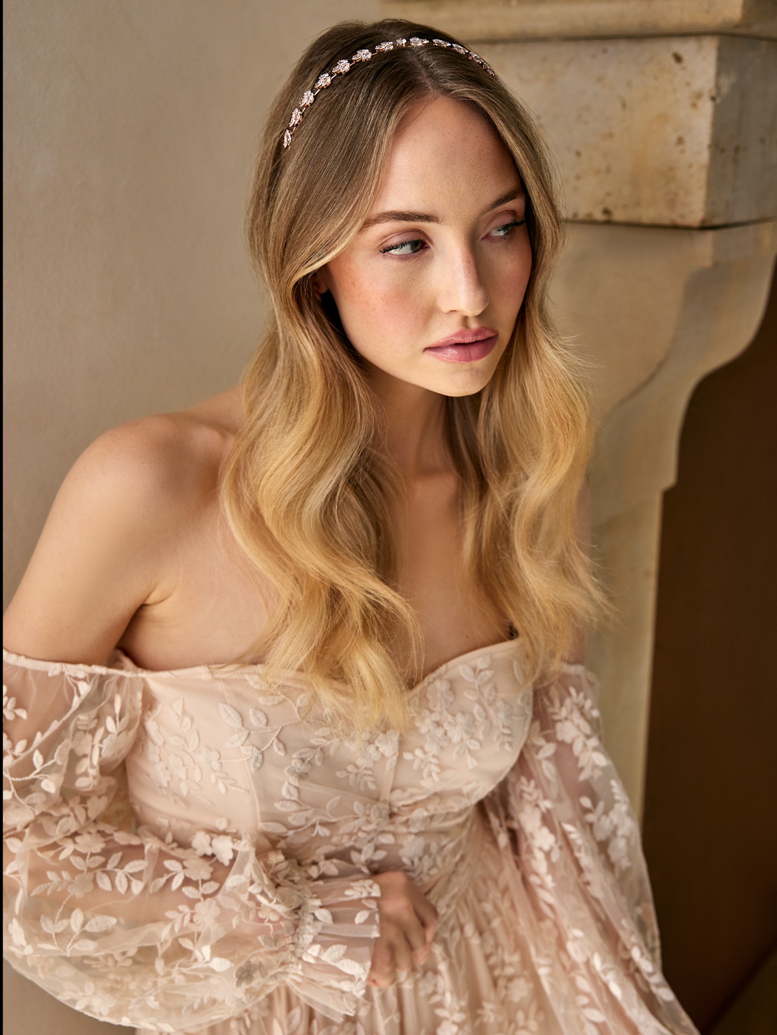 Portret kobiety z blond włosami w sukni z koronkowymi rękawami i ozdobną opaską, pozującej przy ścianie.