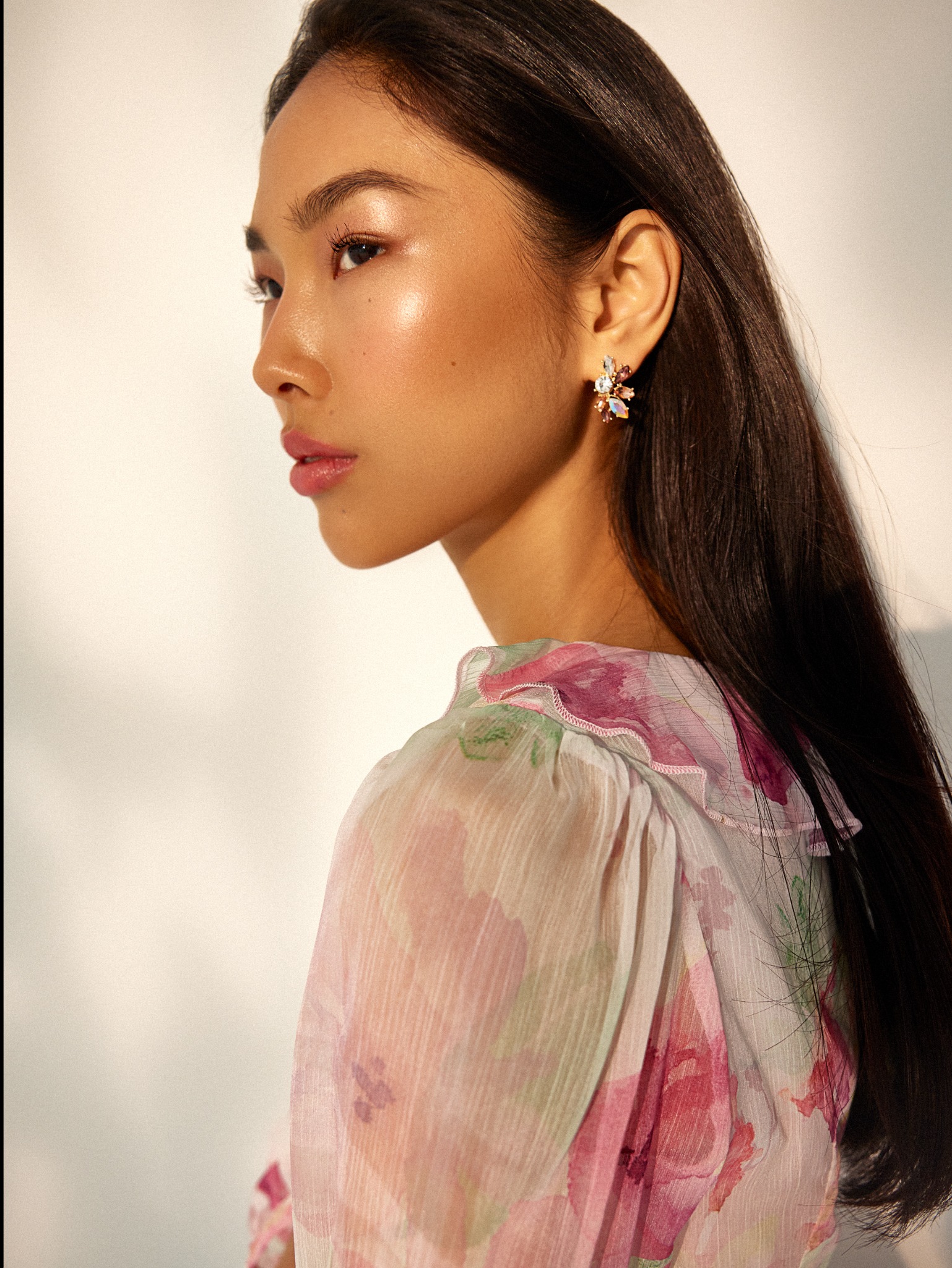 Profil kobiety z błyszczącym makijażem, podkreślonymi ustami i kwiatowymi kolczykami na tle jasnej ściany.