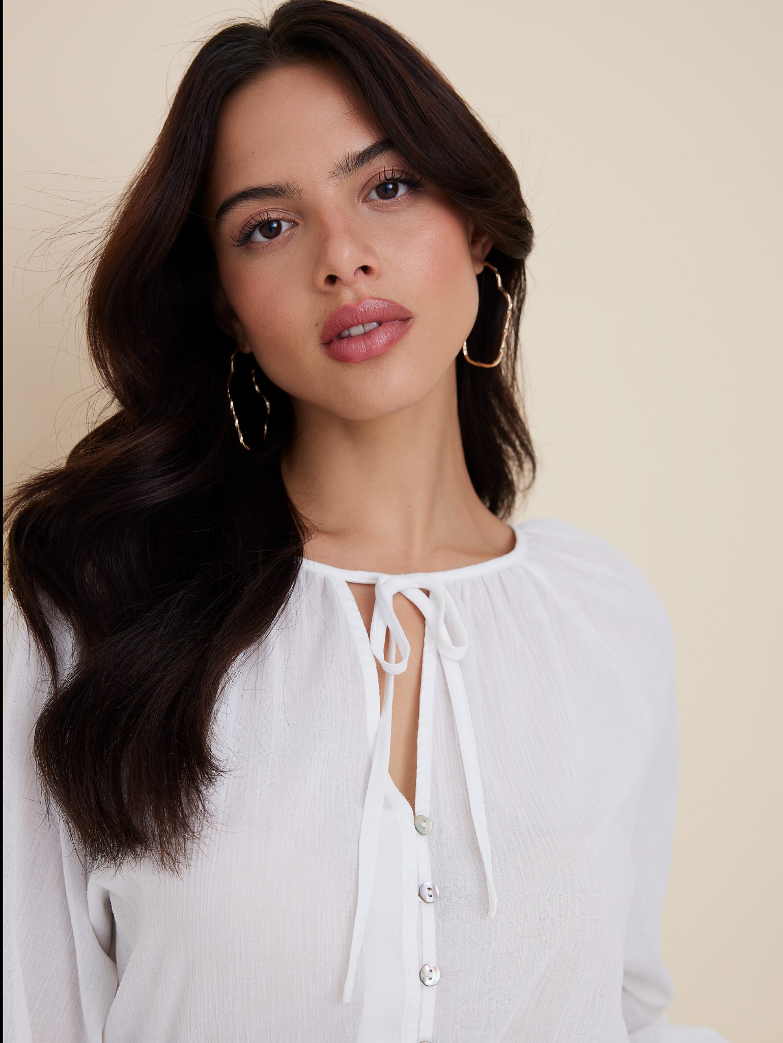 Portret kobiety z subtelnym, naturalnym makijażem, uwydatniającym rysy twarzy, z delikatnie podkreślonymi ustami i brązowymi włosami, noszącej duże, złote kolczyki-koła.