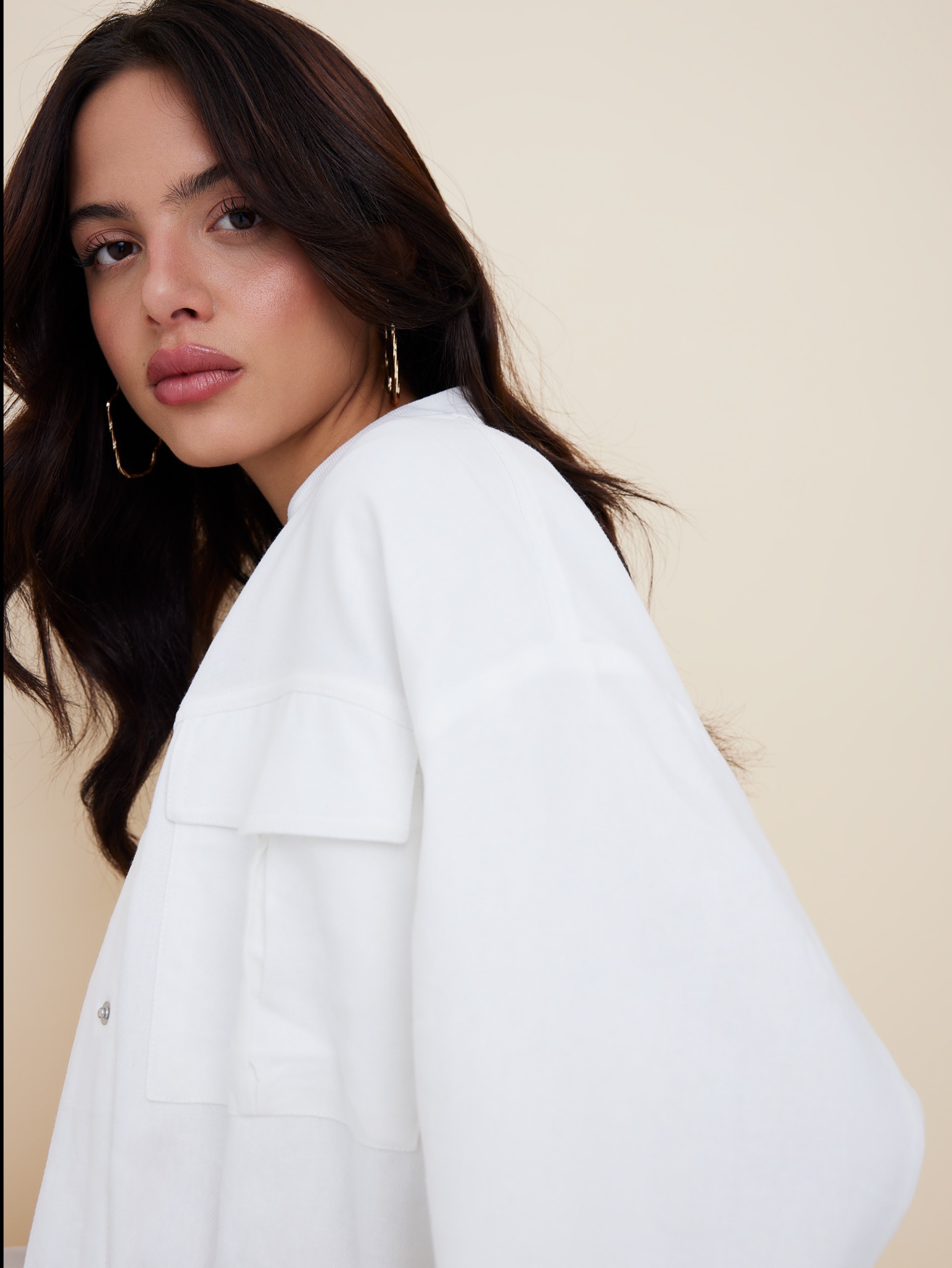 Portret kobiety z delikatnym, błyszczącym makijażem ust i policzków, ciemnymi, falowanymi włosami i złotymi kolczykami na beżowym tle.