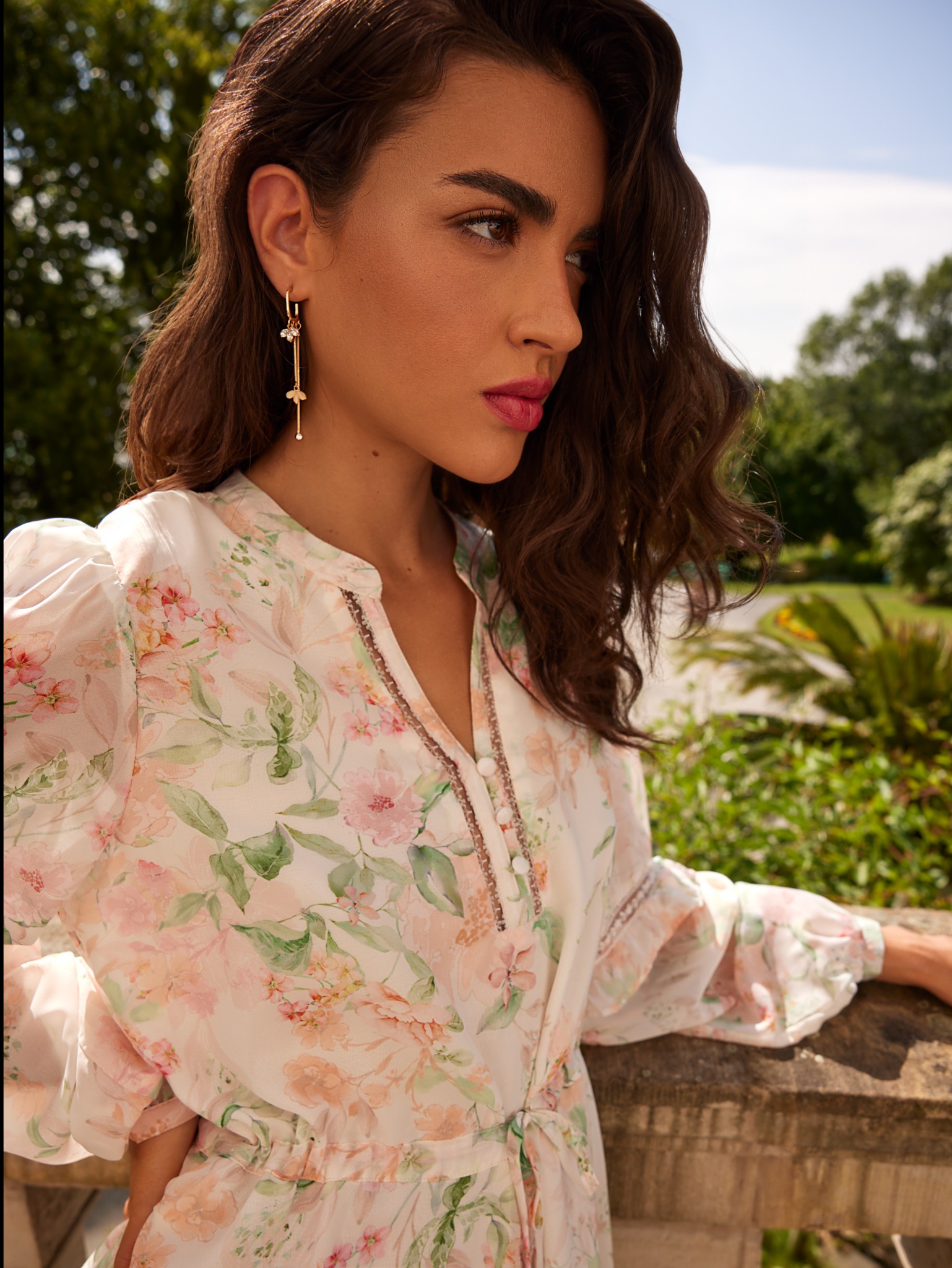 Portret kobiety z delikatnym, różowym makijażem ust i subtelnymi kolczykami w kształcie kwiatów, na tle zieleni ogrodu.