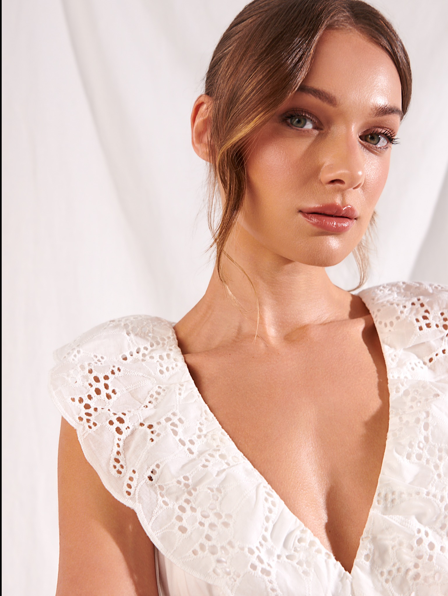 Portret kobiety z subtelnym, rozświetlającym makijażem, podkreślającym zielone oczy i pełne, błyszczące usta, włosy upięte z luźnymi pasmami, ubrana w białą, ażurową sukienkę na tle białej tkaniny.