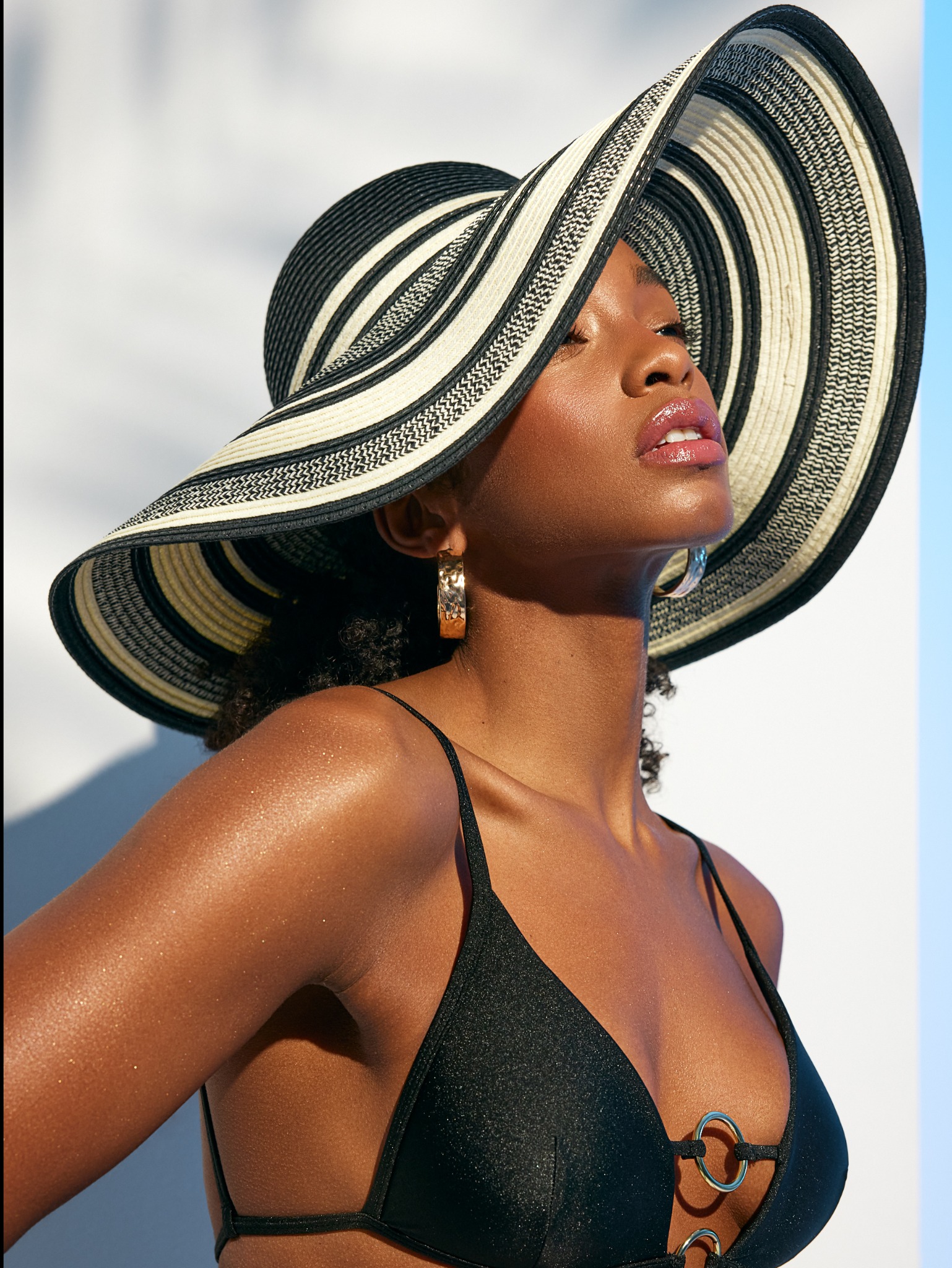 Portret kobiety w czarnym bikini i kapeluszu w paski, z błyszczącym makijażem ust i złotymi kolczykami, spoglądającej w górę w jasnym świetle.
