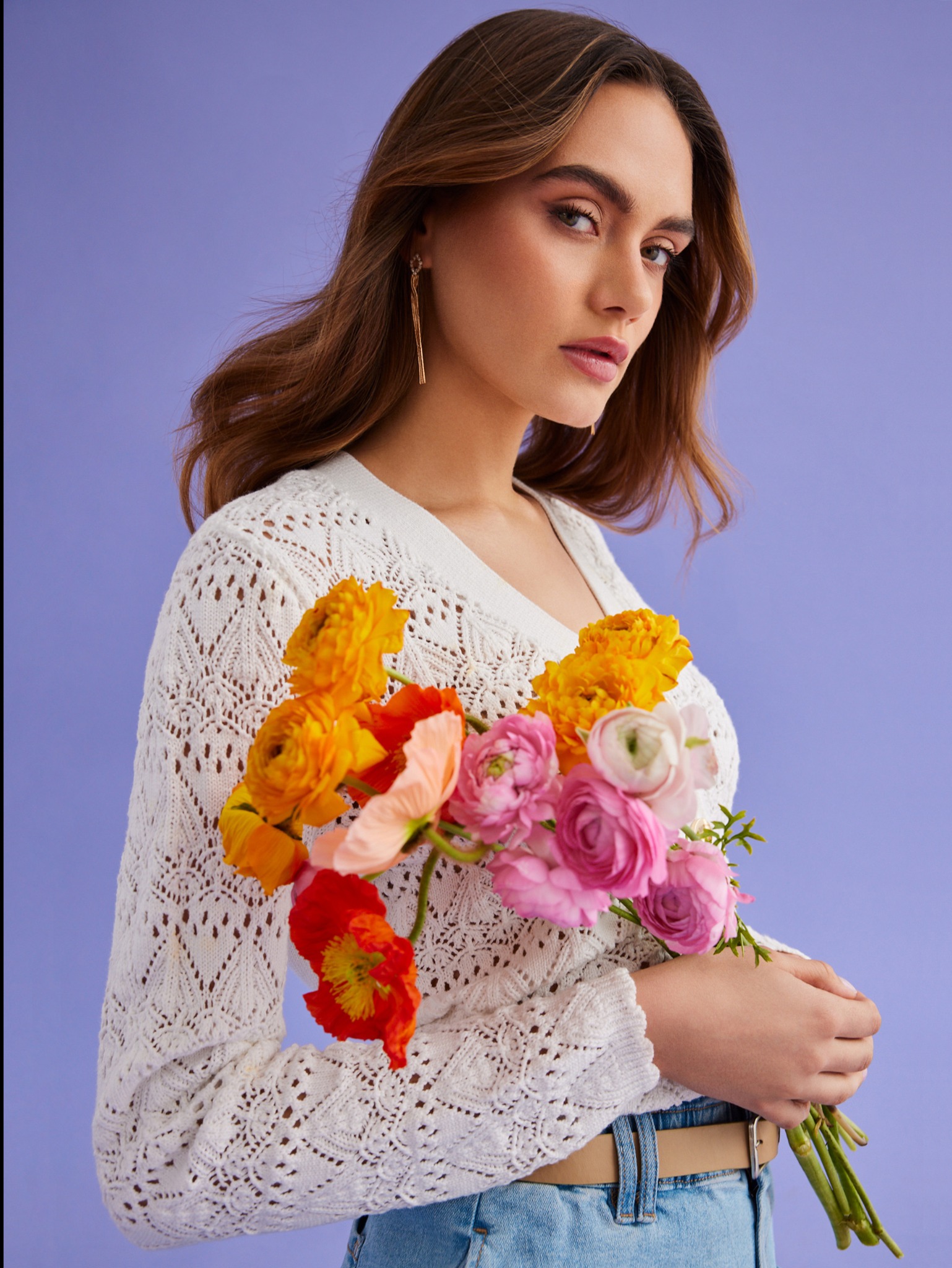 Portret kobiety z delikatnym makijażem, trzymającej bukiet kolorowych kwiatów na fioletowym tle, w białym ażurowym swetrze i dżinsach.
