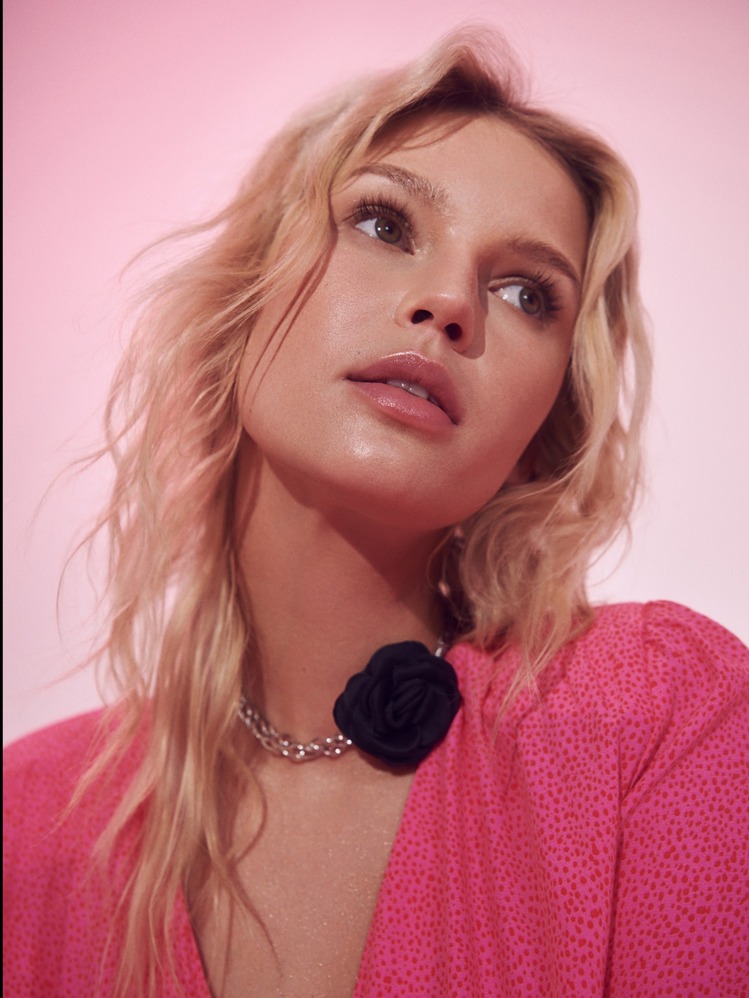 Portret młodej kobiety z blond włosami i delikatnym makijażem, patrzącej w górę, na różowym tle, z widocznym błyszczącym makijażem na ustach i subtelnym cieniem na powiekach.