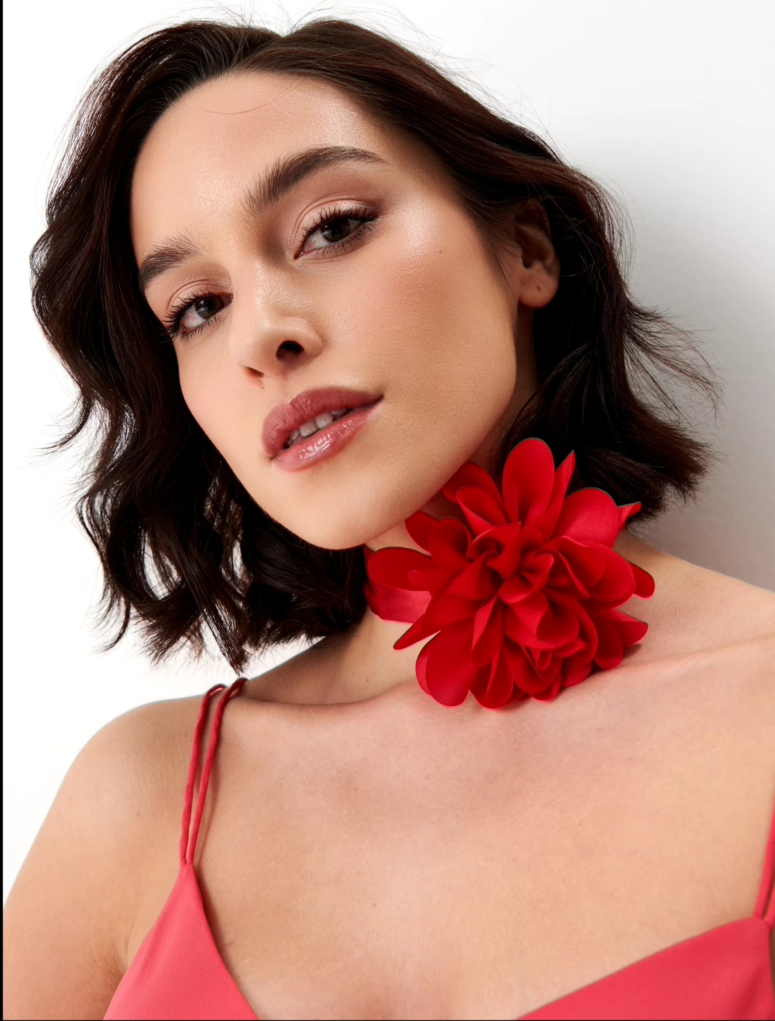 Portret kobiety z delikatnym, naturalnym makijażem, błyszczącymi ustami i subtelnym cieniem do powiek, krótkie, ciemne, falowane włosy, ozdobiona dużą, czerwoną kwiatową broszką przy szyi,...