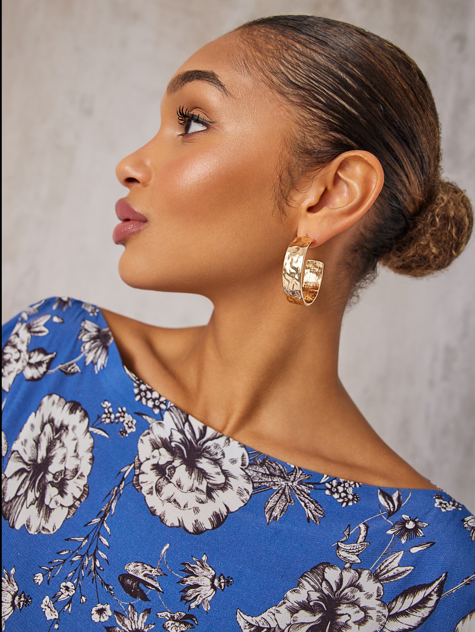 Profil kobiety z delikatnym makijażem, upiętymi włosami i złotymi kolczykami w kształcie koła na tle szarej ściany, ubrana w bluzkę w niebieskie kwiaty.