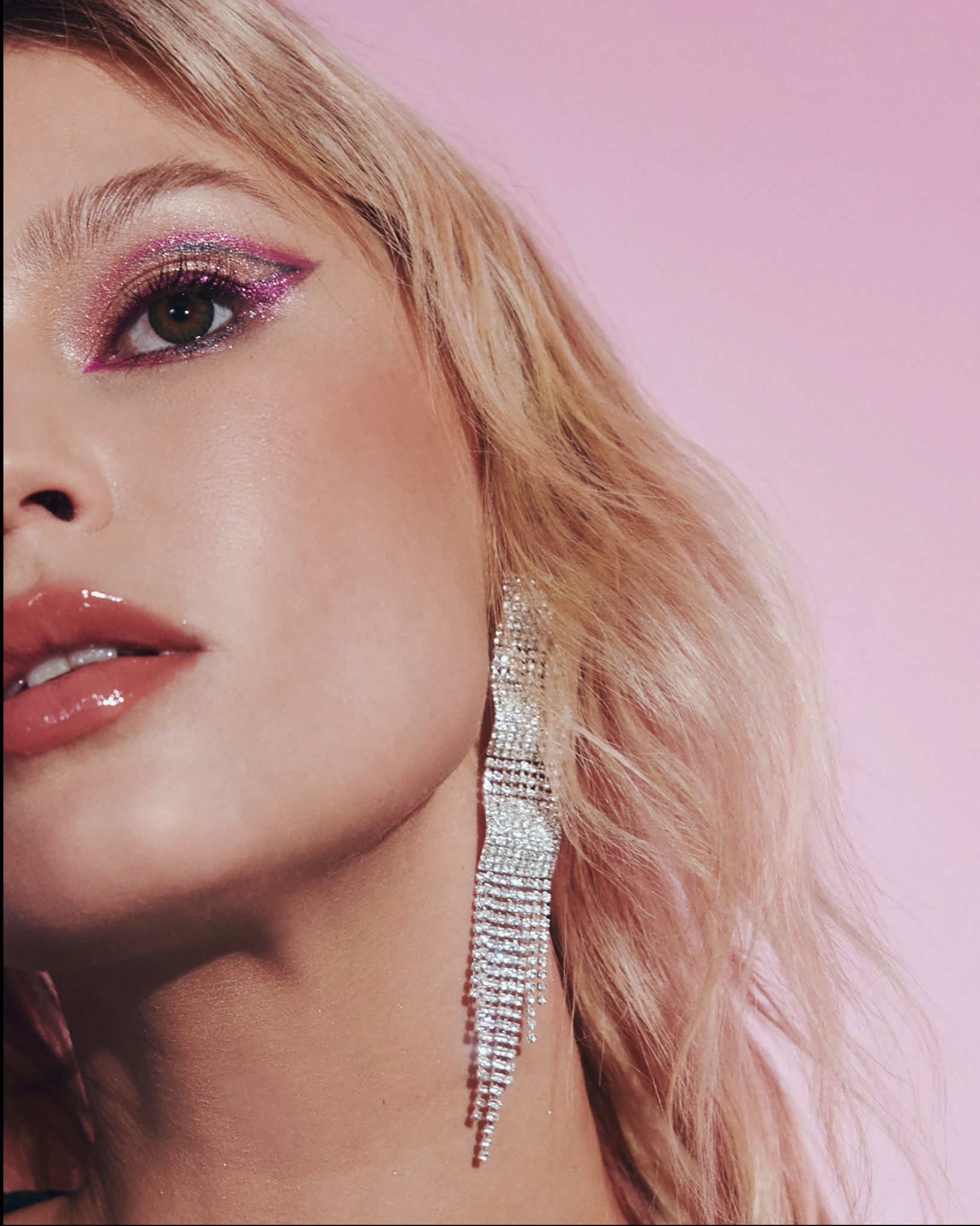 Portret z bliska: Kobieta z błyszczącym, różowo-złotym cieniem na powiekach, pełnymi, błyszczącymi ustami i długim, srebrnym kolczykiem na tle różowego tła.