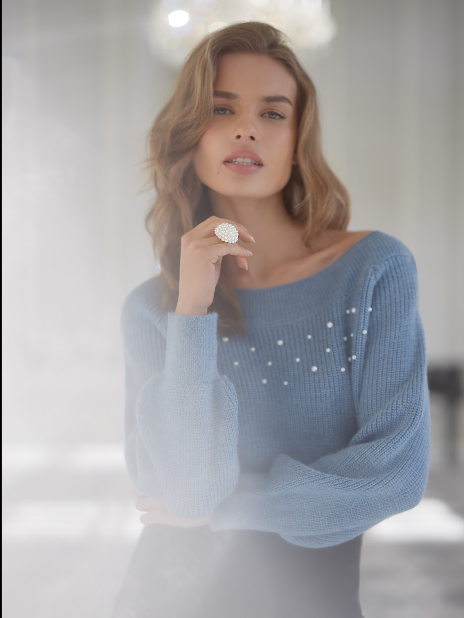 Portret kobiety z delikatnym, naturalnym wyglądem, prezentującej pierścionek z pereł i sweter z perłami, z rozpuszczonymi blond włosami i subtelnym połyskiem na ustach.