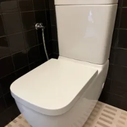 Biała, podwieszana toaleta z chromowanym zaworem kątowym na tle ciemnych płytek i jasnej podłogi w geometryczny wzór. Nowoczesna łazienka po remoncie.