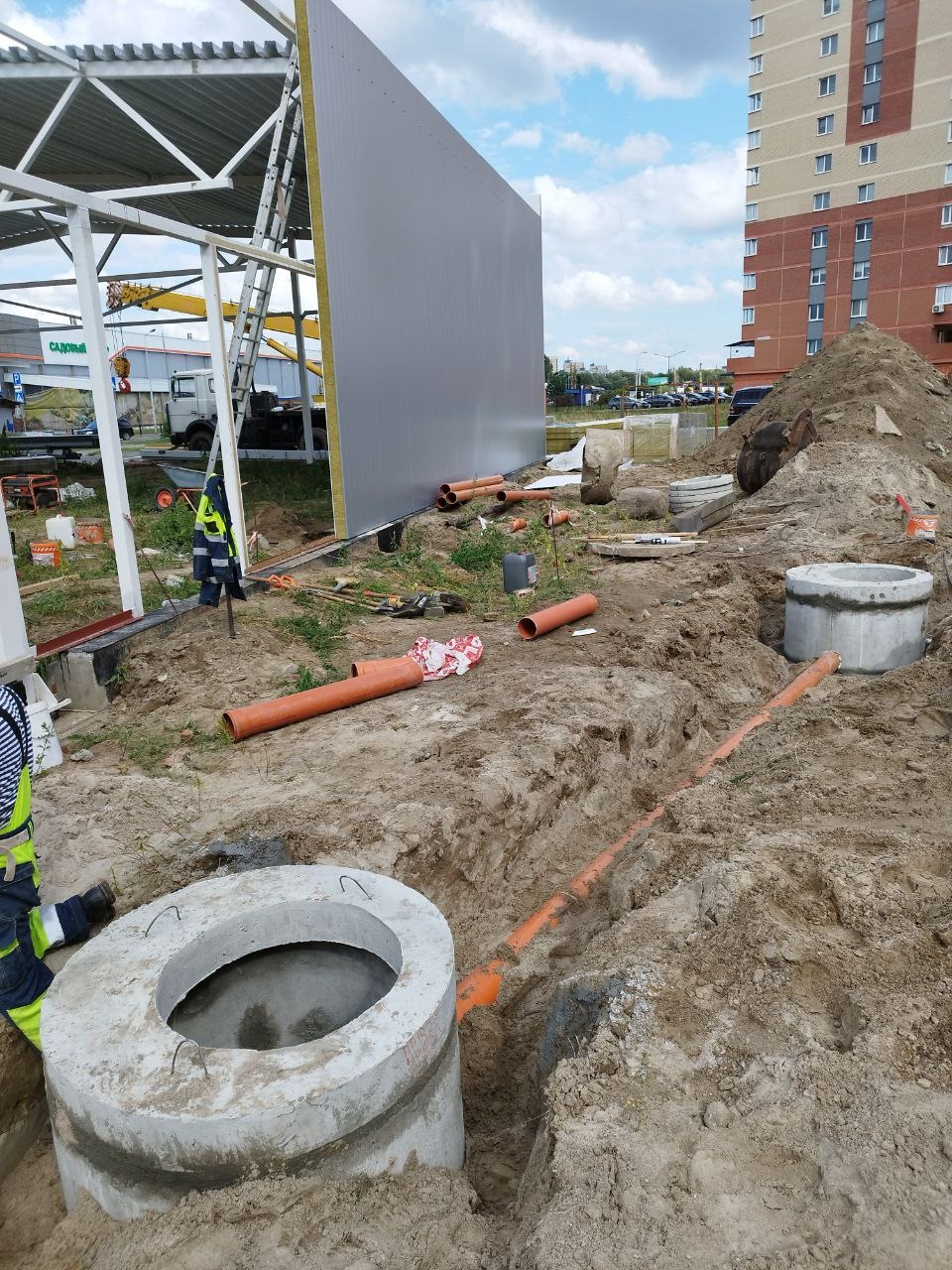 Montaż betonowych studni i rur kanalizacyjnych w wykopie ziemnym. Widoczne narzędzia i fragment budynku w tle. Prace ziemne w trakcie realizacji.