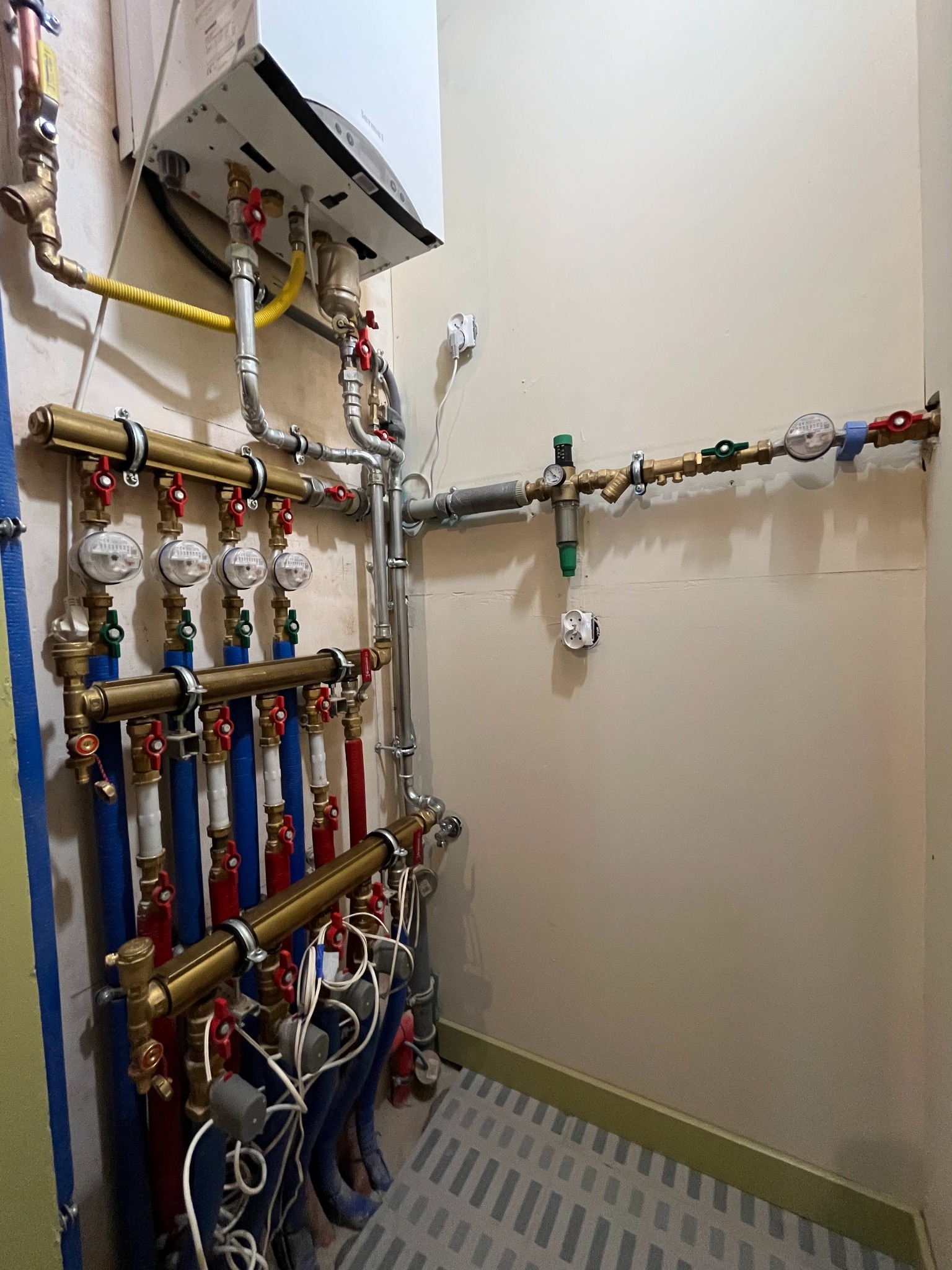 Złożona instalacja CO z piecem gazowym, liczniki wody, rury miedziane i plastikowe z zaworami, szare ściany i dywan w pomieszczeniu technicznym.