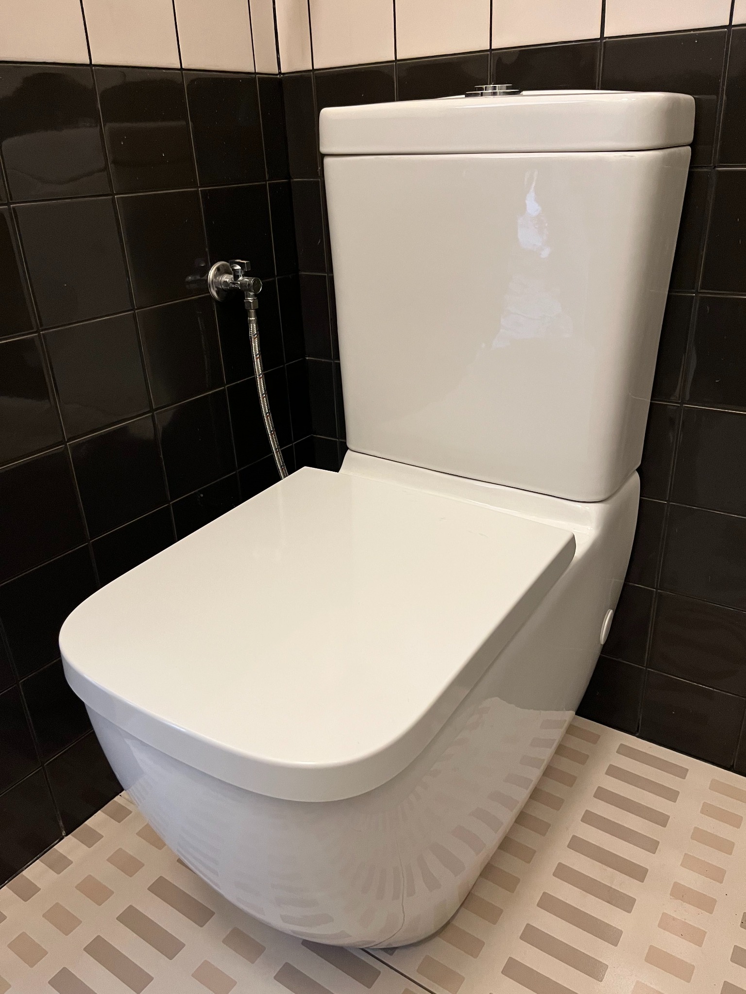 Biała, podwieszana toaleta z chromowanym zaworem kątowym na tle ciemnych płytek i jasnej podłogi w geometryczny wzór. Nowoczesna łazienka po remoncie.