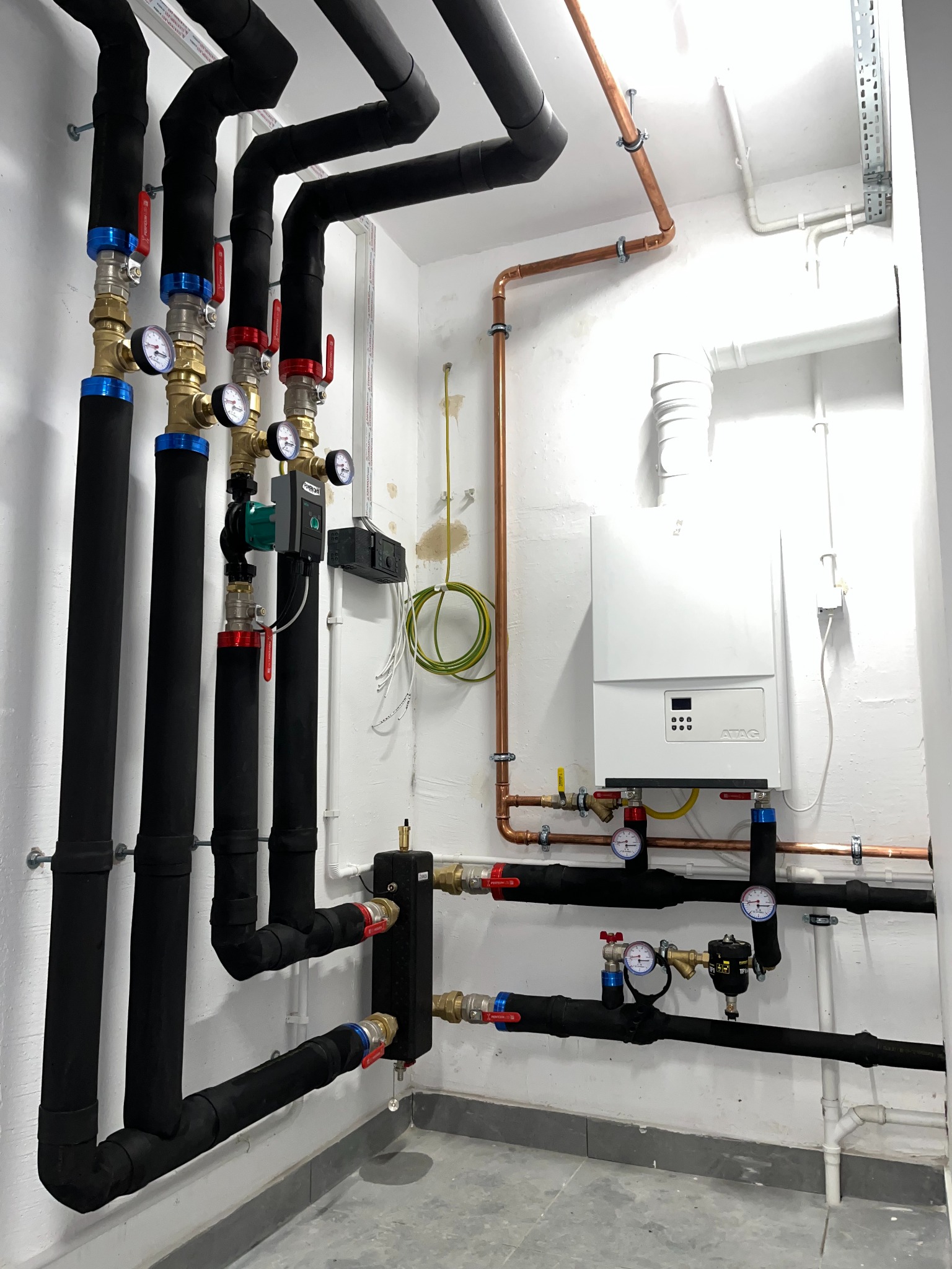Nowoczesna kotłownia z piecem gazowym ATAG, miedzianymi rurami i czarnymi, izolowanymi przewodami. Manometry i zawory z czerwonymi i niebieskimi uchwytami.