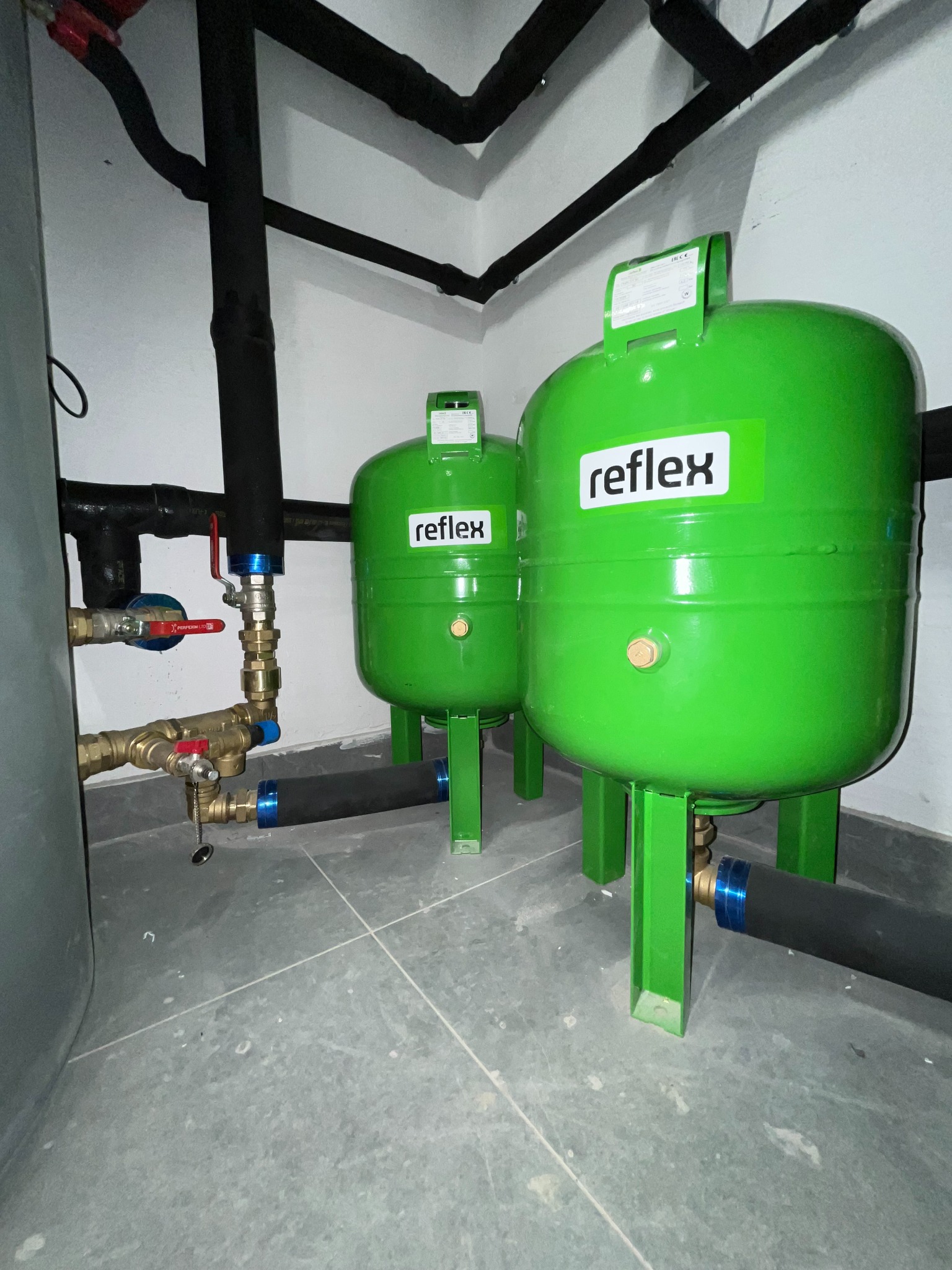 Dwa zielone zbiorniki Reflex z instalacją hydrauliczną i rurami w kotłowni. Widoczne zawory, złączki i czarne rury. Szare płytki na podłodze.