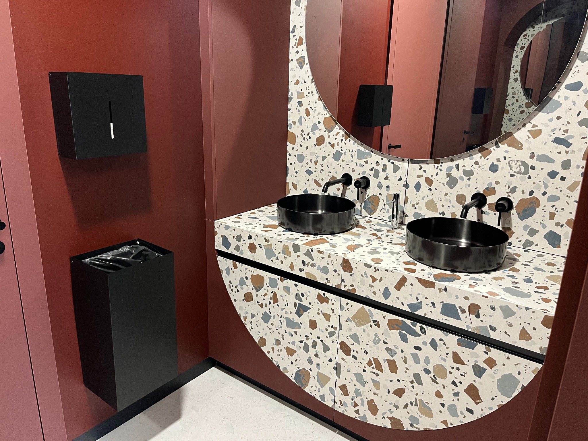 Nowoczesna łazienka w centrum biznesowym: dwie czarne umywalki na blacie z lastryko, okrągłe lustro, dozownik mydła i kosz na śmieci w kolorze ścian.
