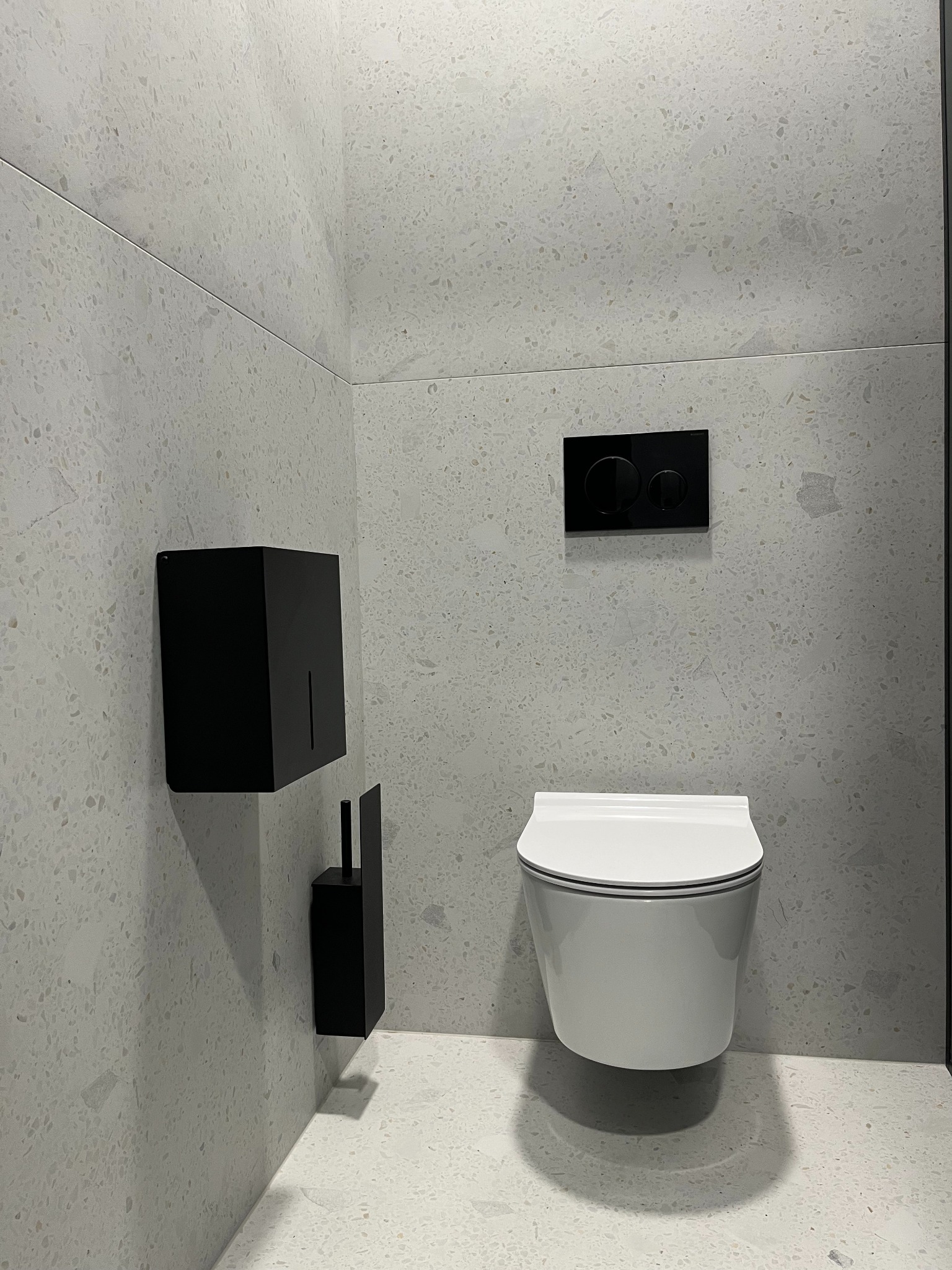 Nowoczesna łazienka z podwieszaną toaletą, czarną armaturą i wykończeniem ścian w jasnym lastryko. Minimalistyczny design, widok z perspektywy narożnika.