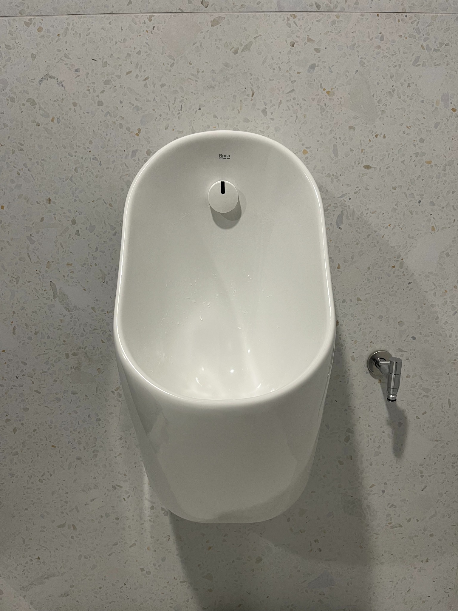 Biały pisuar ceramiczny zamontowany na ścianie z mozaiką w łazience. Widoczny zawór dopływu wody. Ujęcie z bliska, prezentujące detale armatury.