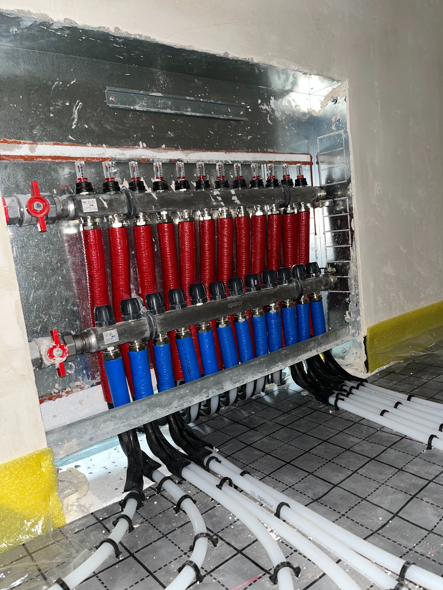 Rozdzielacz obiegów ogrzewania podłogowego Uponor z czerwonymi i niebieskimi rurami, zamontowany w metalowej obudowie, widok na ułożone białe rury na folii z rastrem.