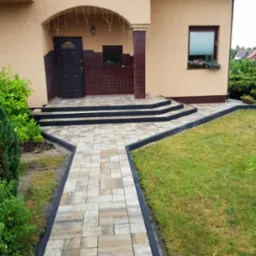 Ułożona ścieżka z kostki brukowej prowadząca do wejścia domu z trzema schodami, obrzeża w kolorze grafitowym, trawnik po prawej stronie, kwiaty w doniczce na parapecie okna.